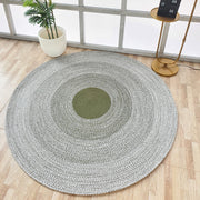 The RAMSHA HOME Handmade Braided Green Color Round Rug BR 079, with a green center and gray rings, rests on a light wood floor beside a potted plant and gold side table, bringing artisanal charm to this airy, window-filled room.
