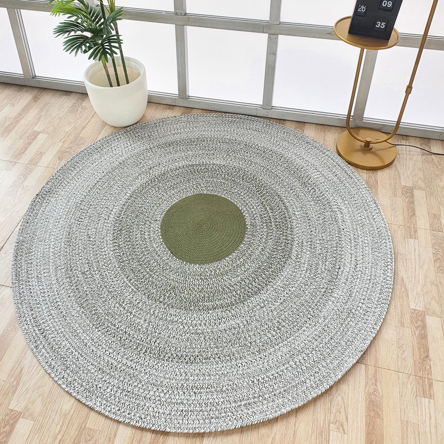 The RAMSHA HOME Handmade Braided Green Color Round Rug BR 079, with a green center and gray rings, rests on a light wood floor beside a potted plant and gold side table, bringing artisanal charm to this airy, window-filled room.