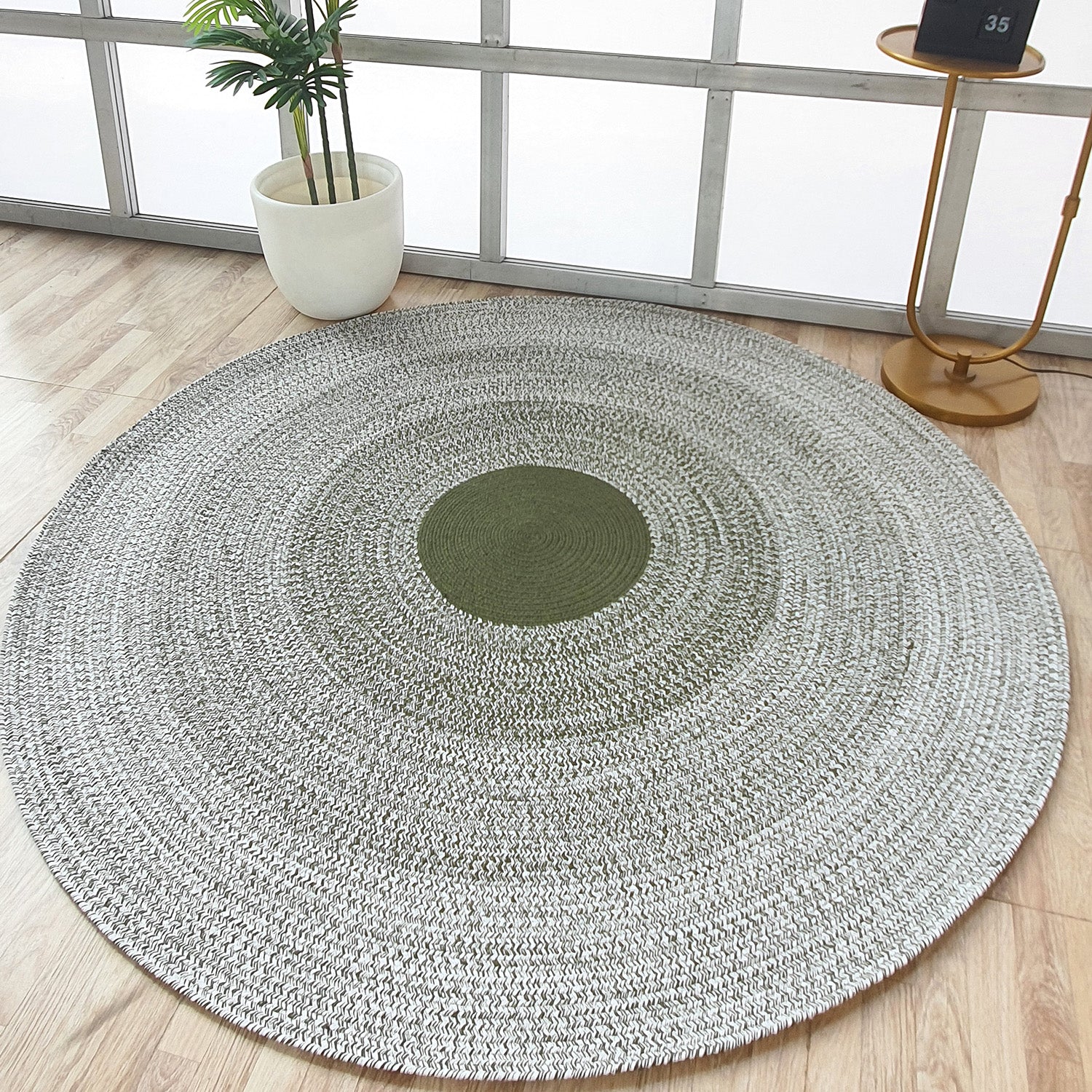 The RAMSHA HOME Handmade Braided Green Color Round Rug BR 079, featuring a green center and gray outer rings, rests on a light wood floor beside a plant and gold table, bringing artisanal charm to this bright, airy room.