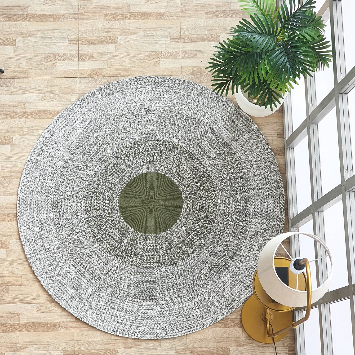 The RAMSHA HOME Handmade Braided Green Color Round Rug BR 079 with green and gray concentric circles rests on a light wood floor near a leafy potted plant, a modern floor lamp, and large windows.