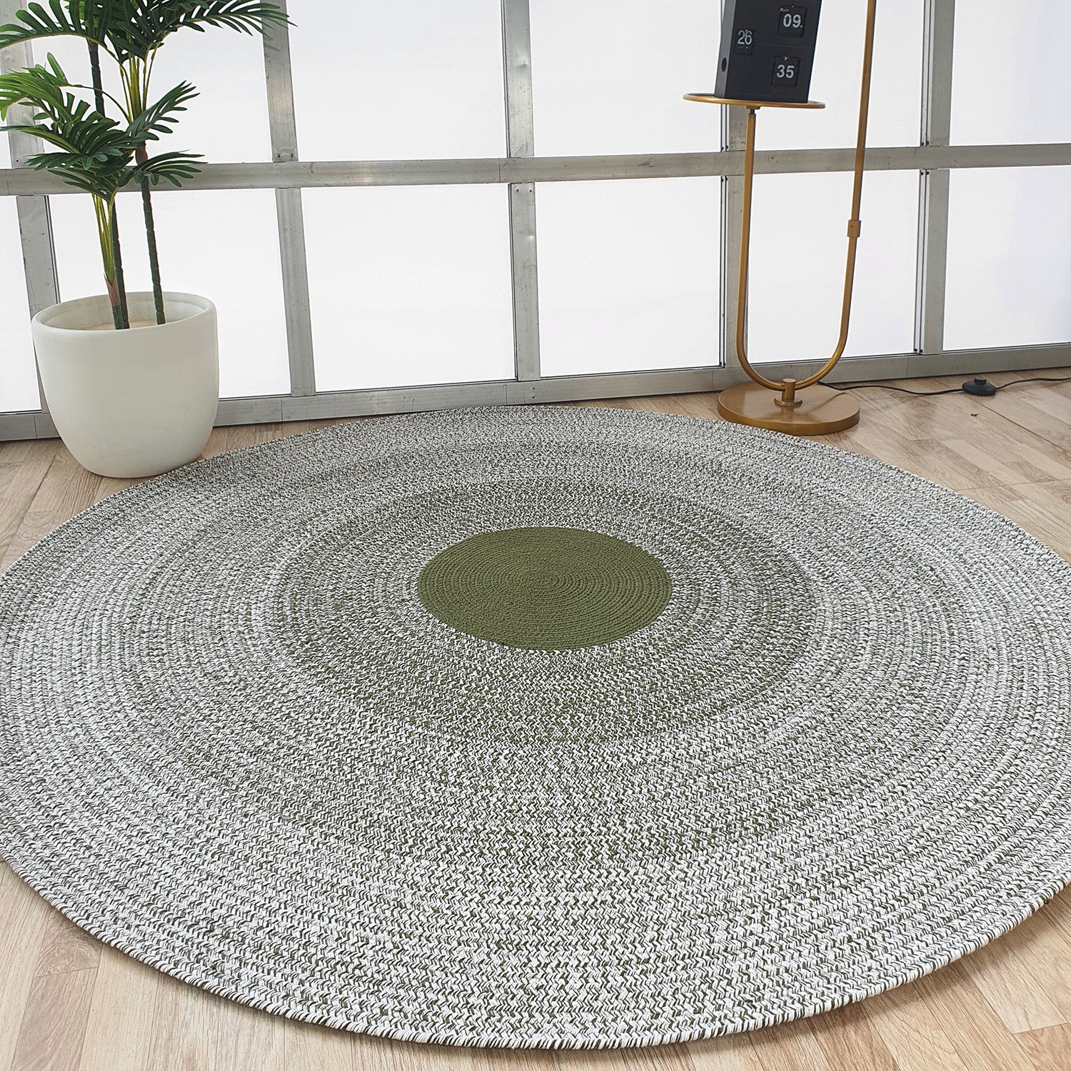 The RAMSHA HOME Handmade Braided Green Color Round Rug BR 079 lies on a light wood floor. Nearby are a potted plant, a floor lamp, and a small black and white calendar by a metal-framed window.