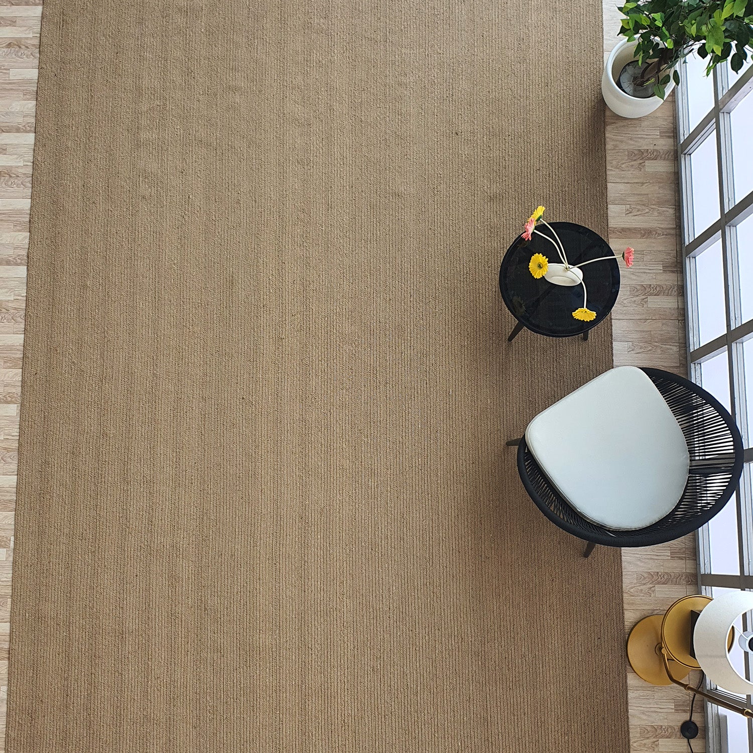 A modern, minimal room features the RAMSHA HOME Natural Color Dining Room Jute Area Braided Rug (BR 081), a black round table with a vase of colorful flowers, a black and white chair, a green potted plant, and large sunlit windows.