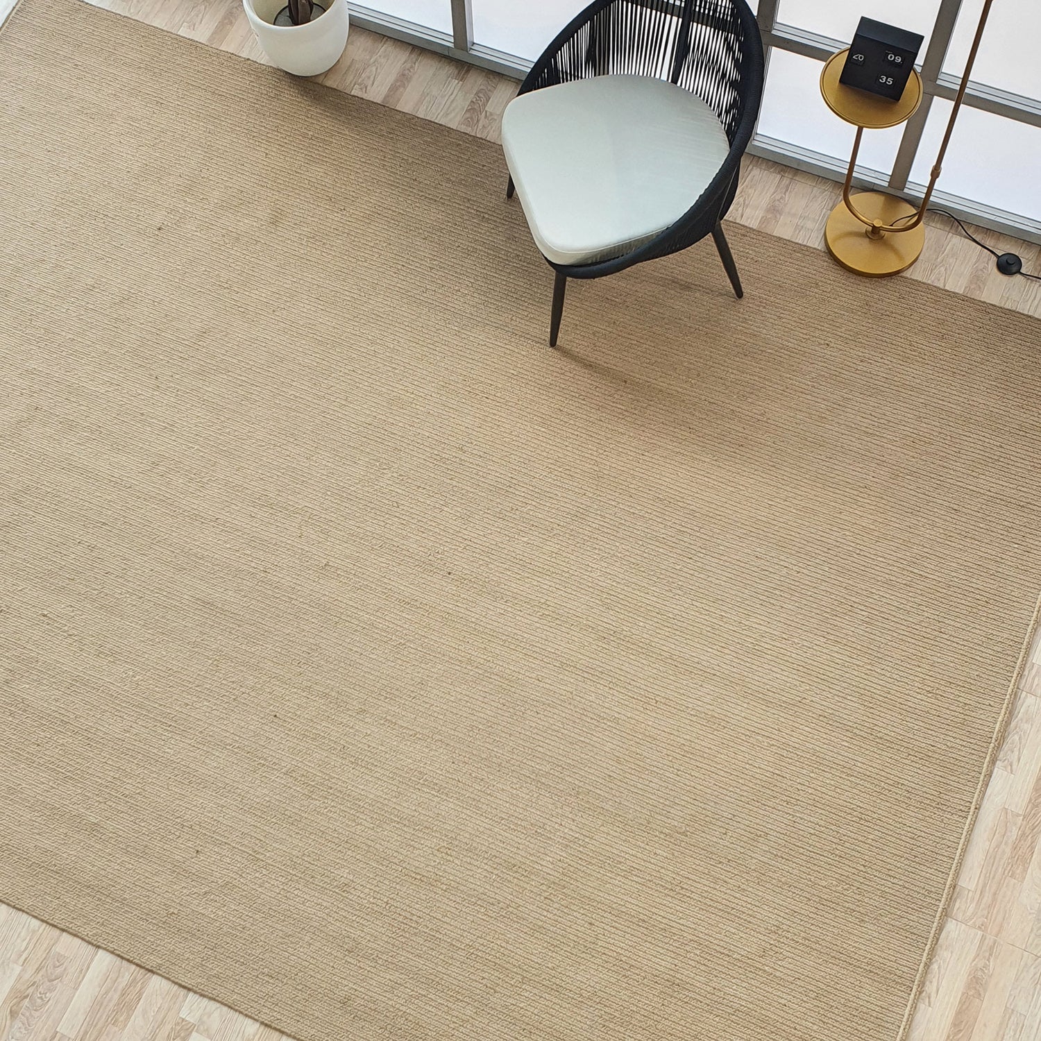 The RAMSHA HOME Natural Color Dining Room Jute Area Braided Rug - BR 081 covers the wood floor. A modern chair, plant, and a gold side table with a flip clock are visible.