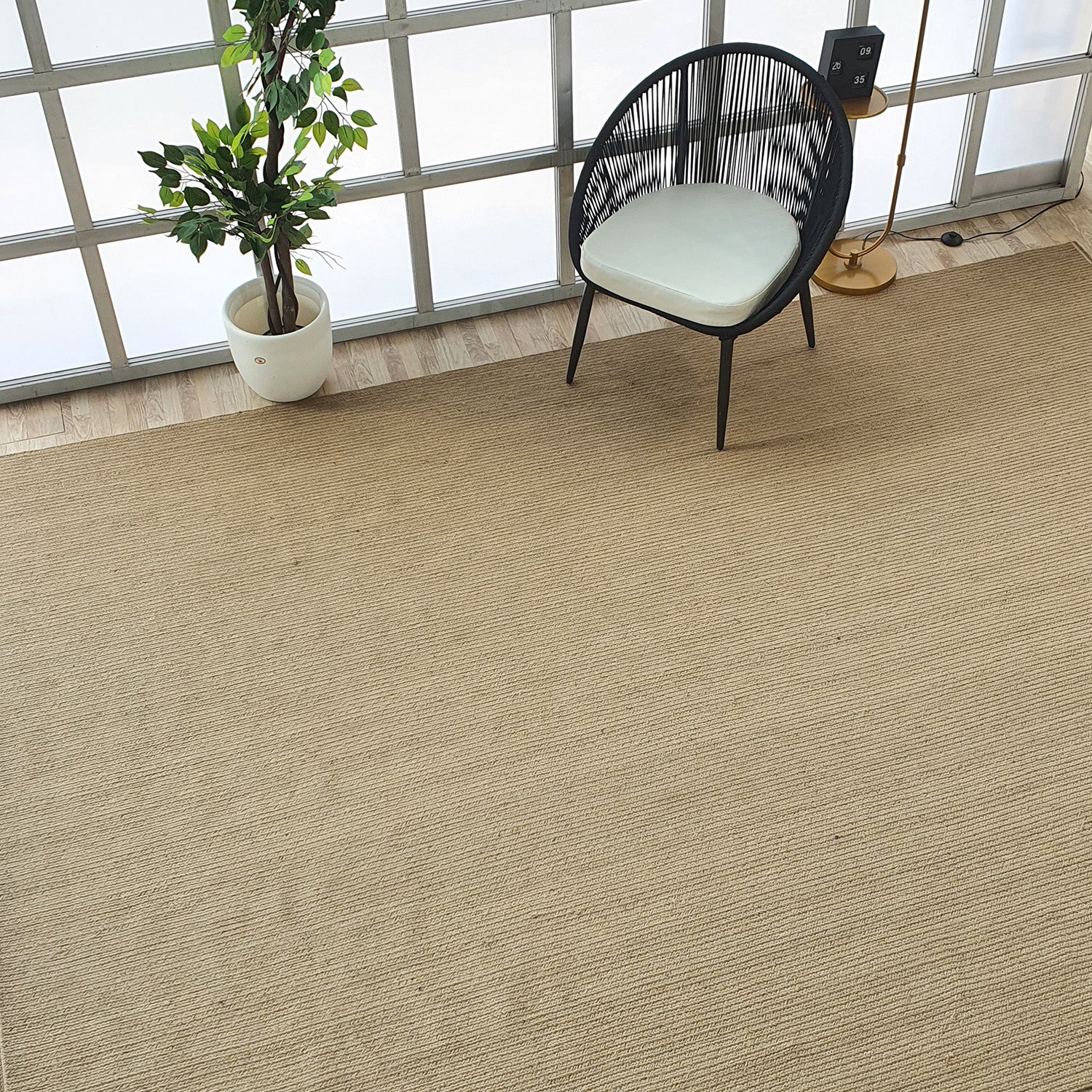 A modern room with a RAMSHA HOME Natural Color Dining Room Jute Area Braided Rug (BR 081), a black wicker chair with white cushion, potted plant, gold floor lamp, and large windows with frosted glass panels.
