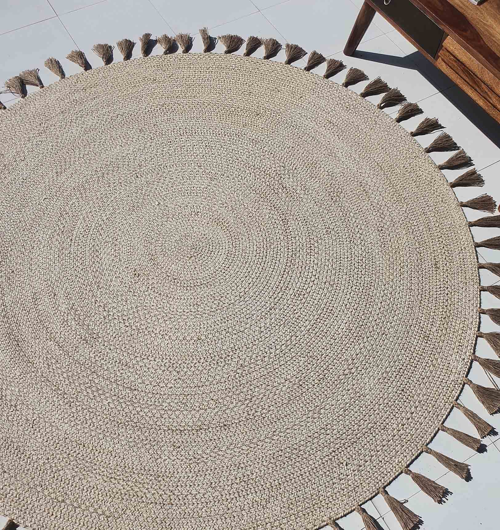The RAMSHA HOME Natural Color Dining Room Jute Area Braided Rug - BR 082, featuring round shape and tassels, is placed on a tiled floor with part of a wooden table or bench visible in the upper right corner.