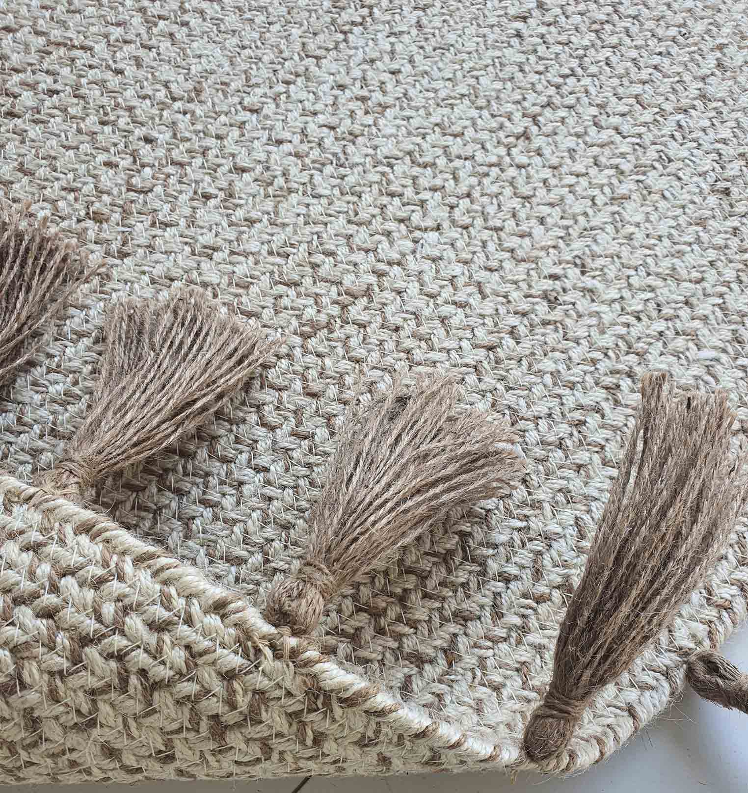 A close-up of the RAMSHA HOME Natural Color Dining Room Jute Area Braided Rug - BR 082 shows its woven texture and four brown tassels on the edge, with visible fiber patterns and a corner slightly folded.
