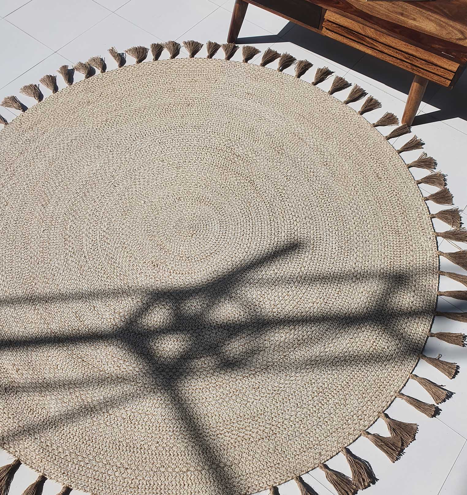 The RAMSHA HOME Natural Color Dining Room Jute Area Braided Rug - BR 082, featuring tasseled edges, lies on white tiles as sunlight highlights its braided texture. A wooden table appears partially in the top right corner.