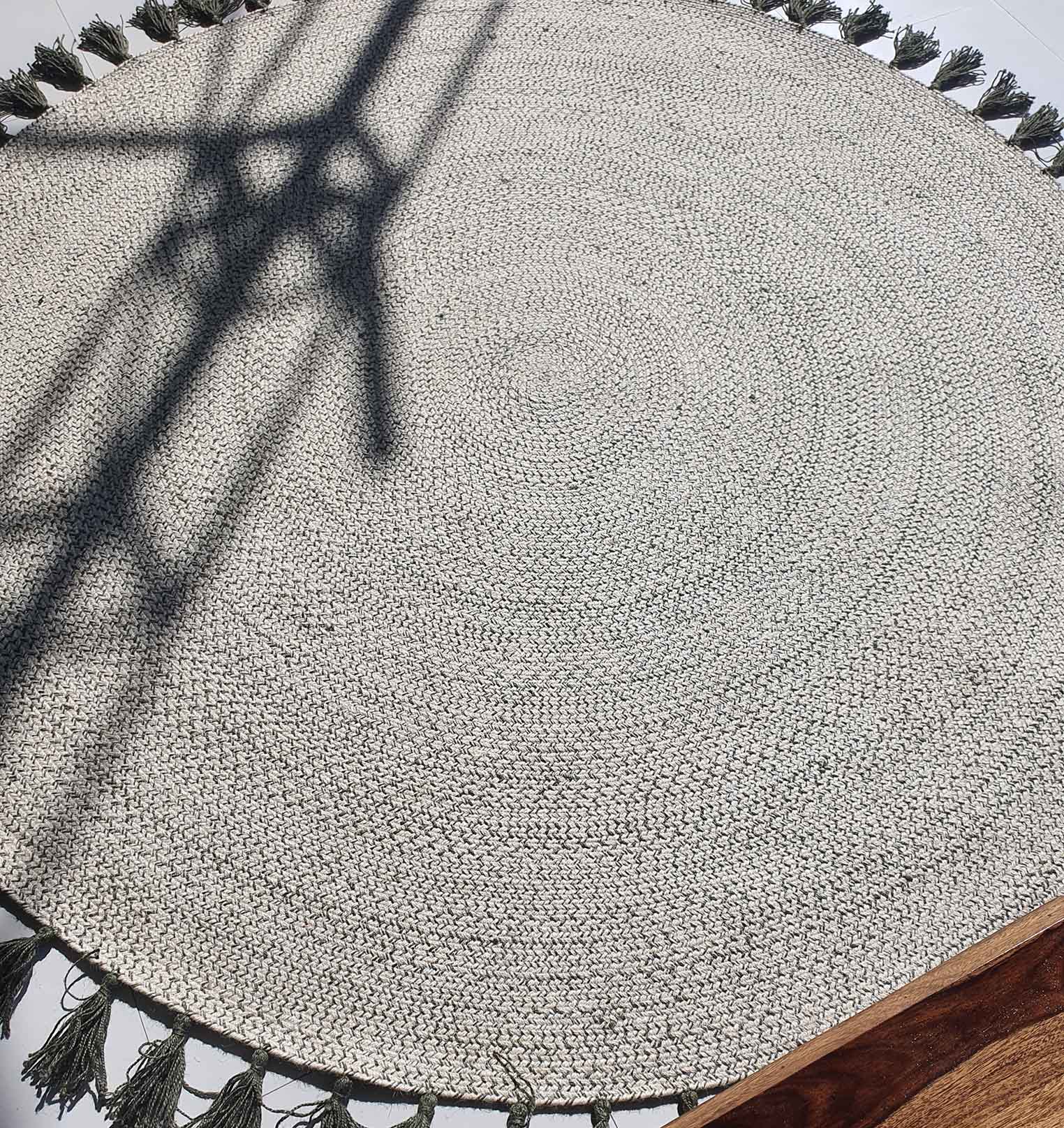 The Amazing Handmade Jute Round Braided Rug with Tassel - BR 083 by RAMSHA HOME is shown in light gray with braided concentric circles and tassels; a metal shadow crosses it, and a wooden surface appears in the lower right corner.