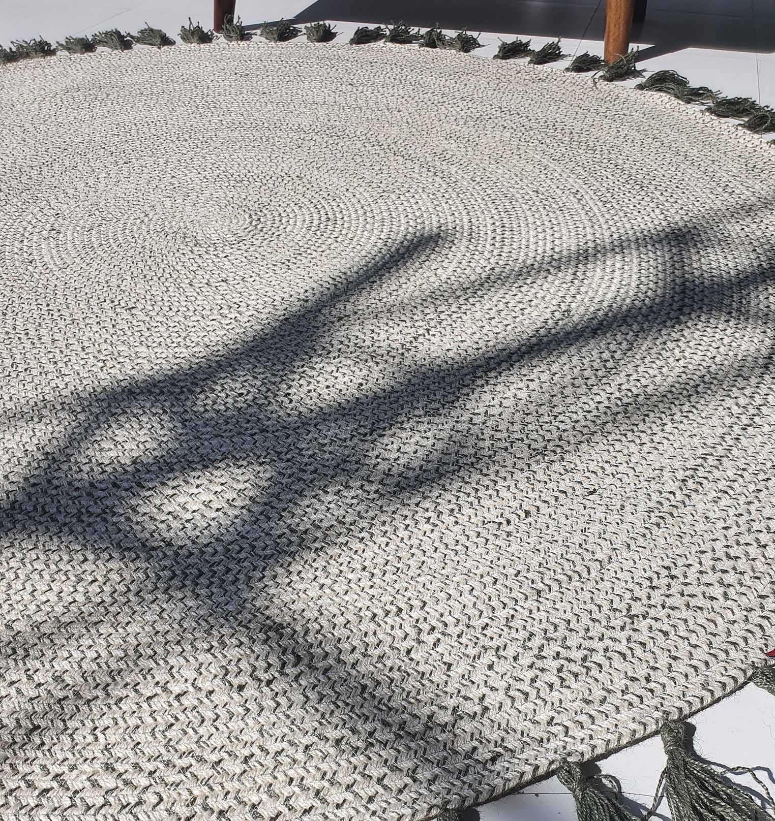 The Amazing Handmade Jute Round Braided Rug with Tassel - BR 083 by RAMSHA HOME sits on a light floor, its braided texture and tasseled edge highlighted by leafy shadows. Two wooden furniture legs appear in the top right corner.