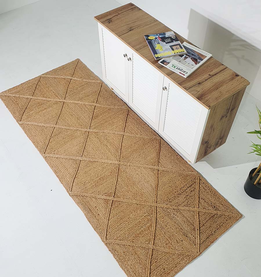 The RAMSHA HOME Amazing Handmade Jute Rug Living Room - BR 084, featuring a tan geometric diamond pattern, is placed on a white floor near a wooden cabinet with white doors, decorated with magazines and a potted plant.