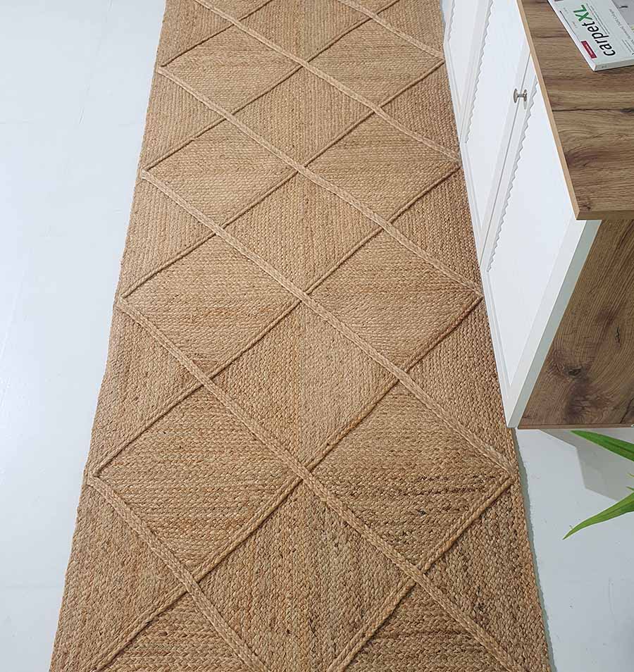 The RAMSHA HOME Amazing Handmade Jute Rug Living Room - BR 084, a long rectangular rug with a diamond pattern, rests on a white floor beside a white cabinet with a wooden top. A green plant is partly visible in the bottom right corner.