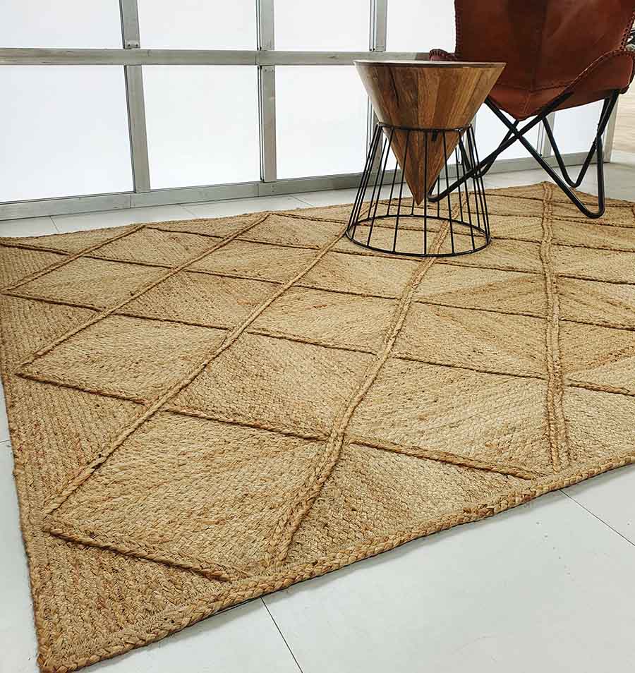 The RAMSHA HOME Amazing Handmade Jute Rug Living Room – BR 084 with a diamond pattern rests on a light tiled floor beside a modern leather chair and metal-wire side table in a bright, minimalist space.