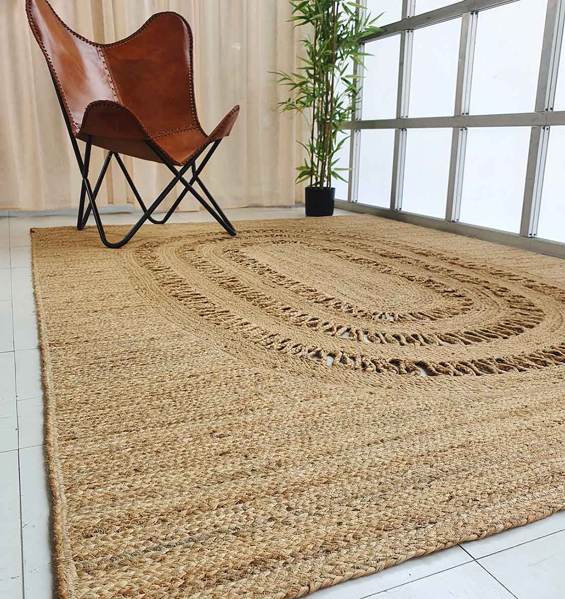 The RAMSHA HOME Exquisite Braided Circle Jute Rug - BR 085 is placed on white tiles beside a brown leather butterfly chair and potted bamboo near large windows with beige curtains in this modern, minimal room.