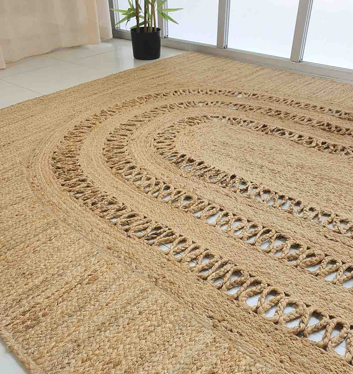 The RAMSHA HOME Exquisite Braided Circle Jute Rug - BR 085 with cut-out patterns decorates a white tile floor beside a potted plant and large windows with beige curtains in a bright room.