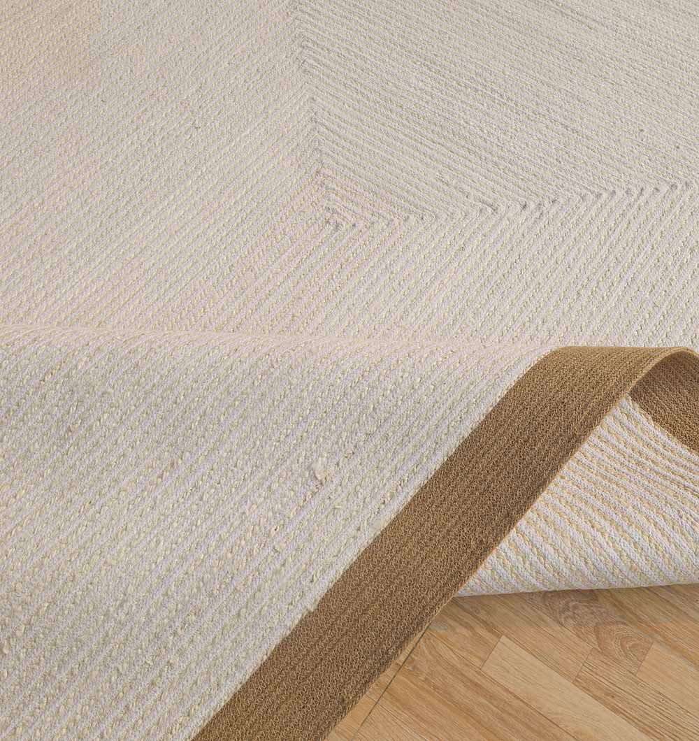A close-up of the RAMSHA HOME Home Decor Rectangle Jute Rug - BR 089 shows its beige textured surface, tan border, and subtle diagonal pattern, with a corner lifted to reveal the wooden floor beneath.
