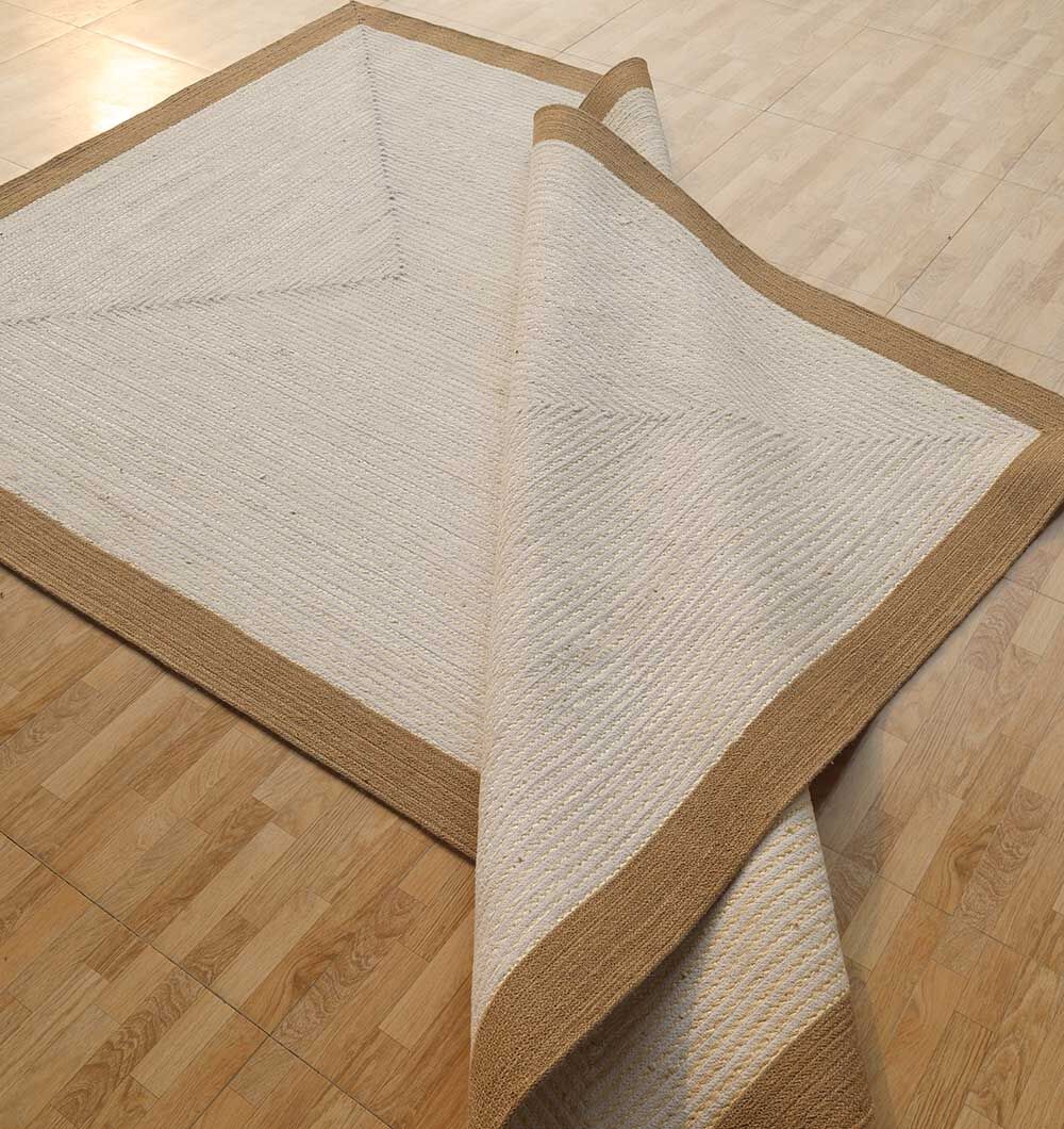 The Home Decor Rectangle Jute Rug - BR 089 by RAMSHA HOME, featuring a beige center and brown border, is partially folded over on a light wood floor.