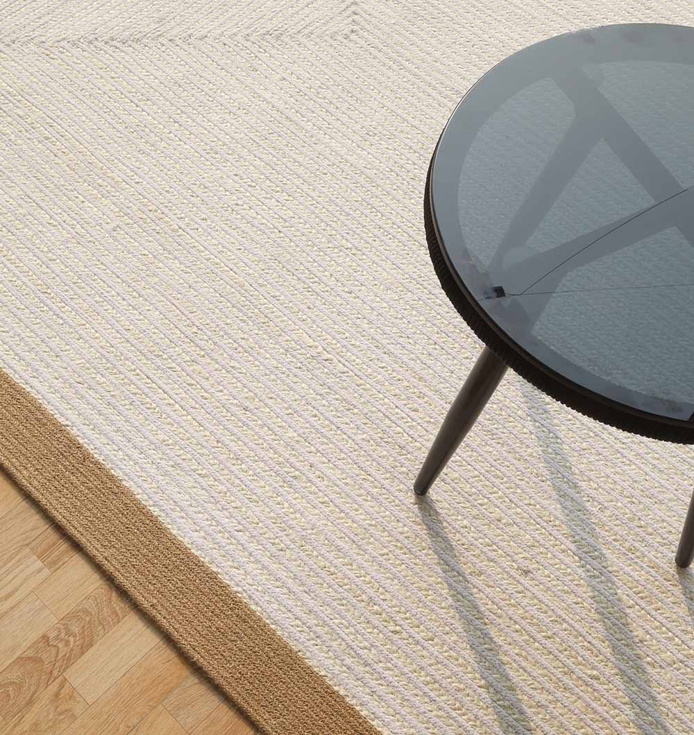 A round glass-top table with black legs sits on the RAMSHA HOME Home Decor Rectangle Jute Rug - BR 089, a textured two-tone jute rug with a tan border, placed on a wooden floor.