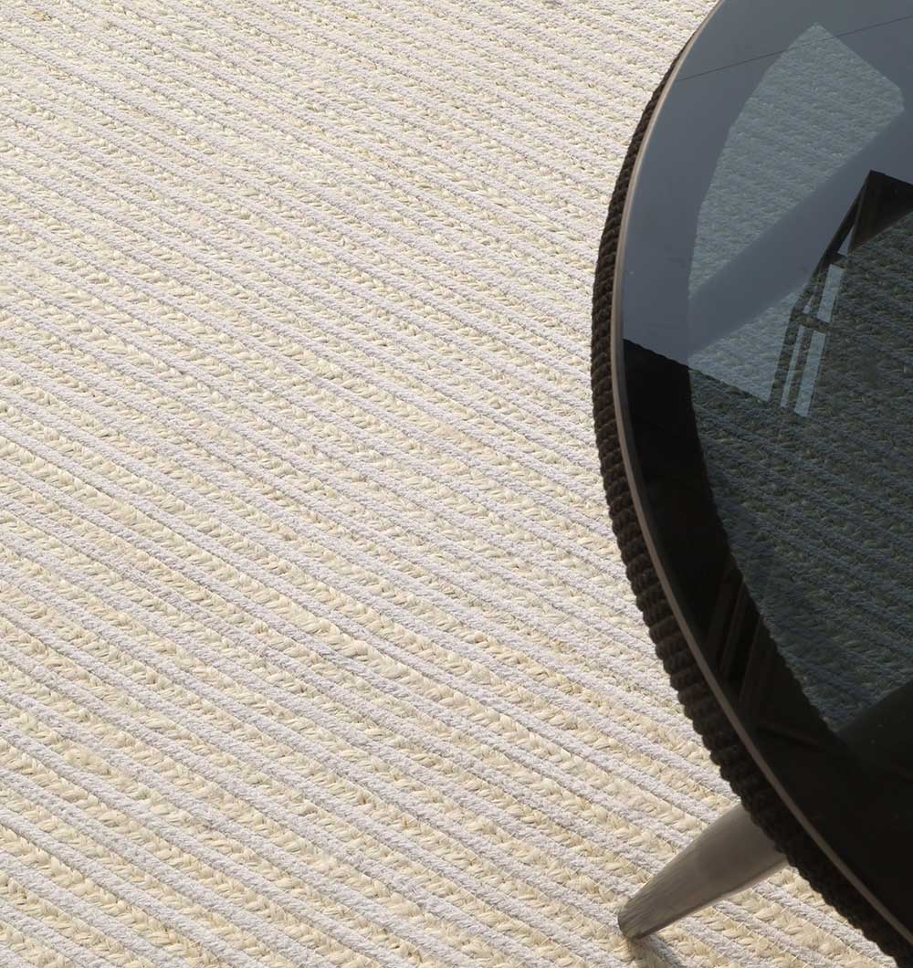 A round glass-top table with metal legs sits on the RAMSHA HOME Home Decor Rectangle Jute Rug - BR 089, featuring a textured, two-tone design with diagonal patterns. The table reflects part of a window and ceiling.