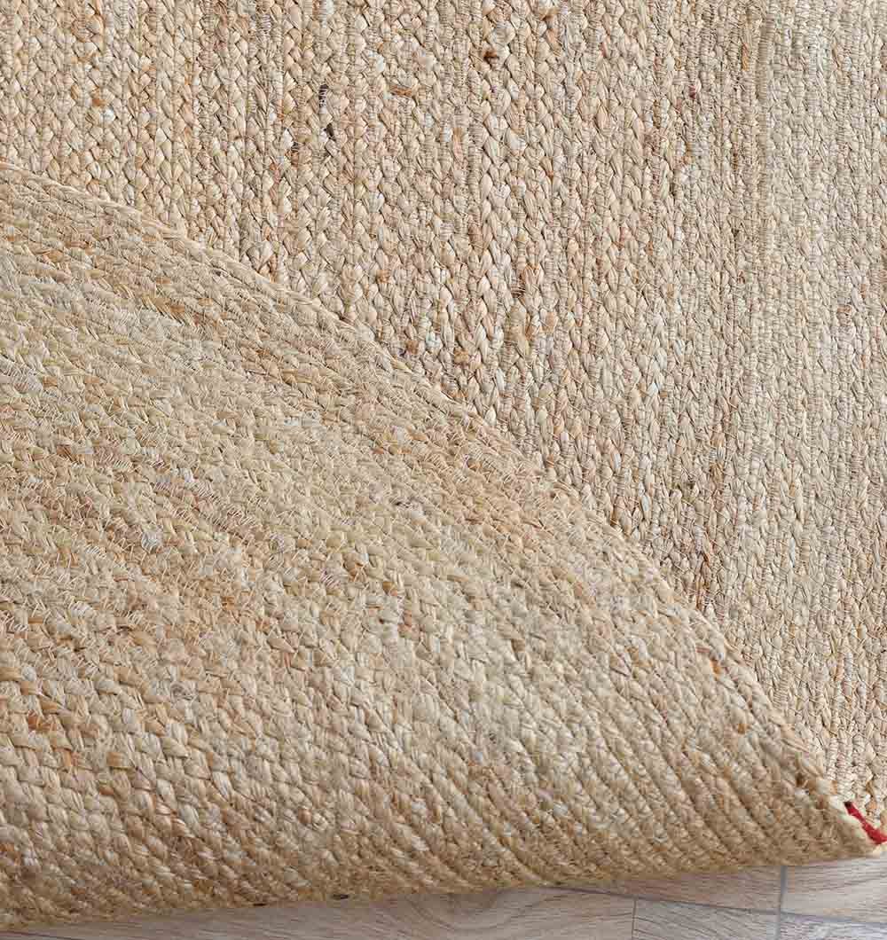 A close-up of the RAMSHA HOME Natural Color Shaped Braided Outdoor Rug 170X240 CM (BR 097), one corner lifted to reveal a light wooden floor, showcases its textured beige jute weave and highlights the rug’s natural beauty.