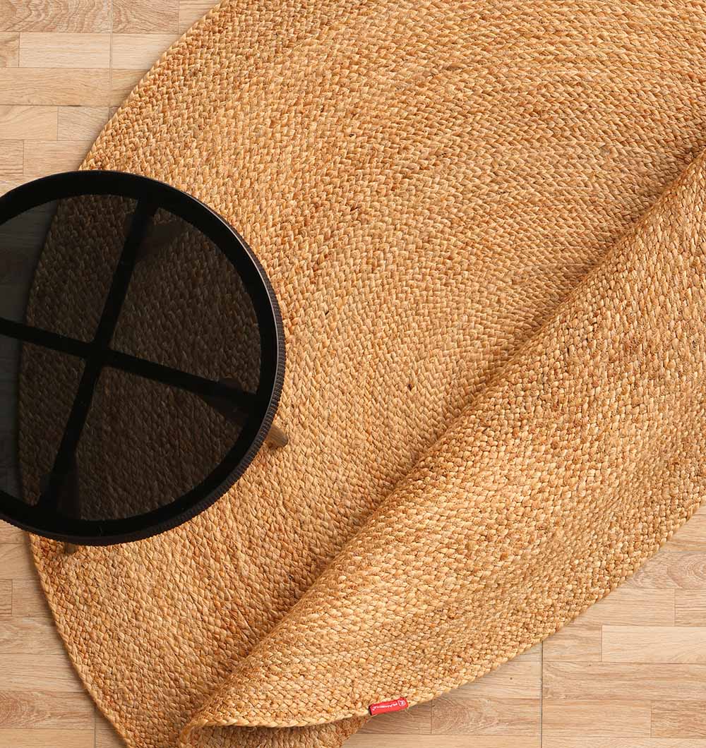 The RAMSHA HOME Round Jute Rug Natural Color - BR 098, partially rolled on a light wood floor, is topped with a black round side table featuring a cross design.