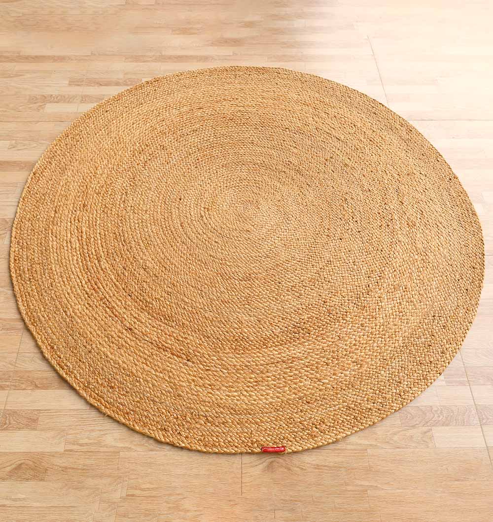 The RAMSHA HOME Round Jute Rug Natural Color - BR 098, featuring a braided concentric pattern and a small red edge tag, lies on a light wooden floor.