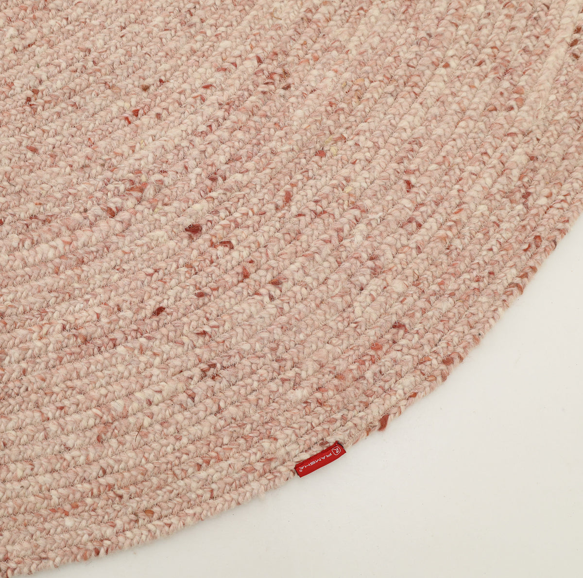 A close-up of the RAMSHA HOME Soft Pink Braided Wool Area Rug Handmade Circular Rug- BR 099 on a white surface, showcasing its textured braided details and a small red tag with white text near the edge. This minimalist circular rug emphasizes its intricate craftsmanship and subtle elegance.