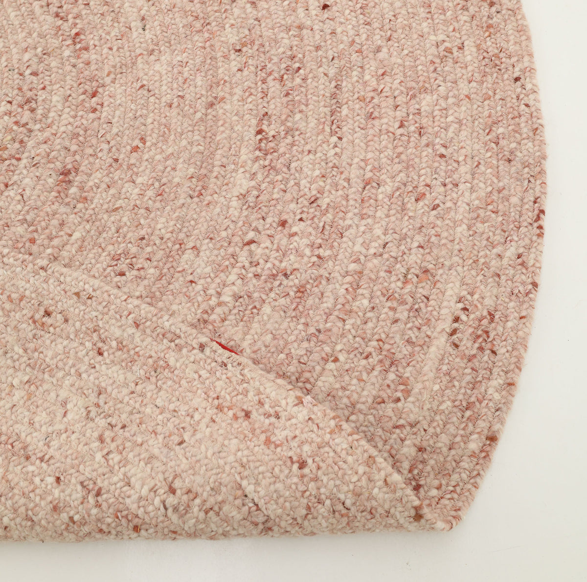 A close-up of the RAMSHA HOME Soft Pink Braided Wool Area Rug (Handmade Circular Rug- BR 099), featuring visible wool loops and subtle speckles, slightly folded at the edge, set against a white background.