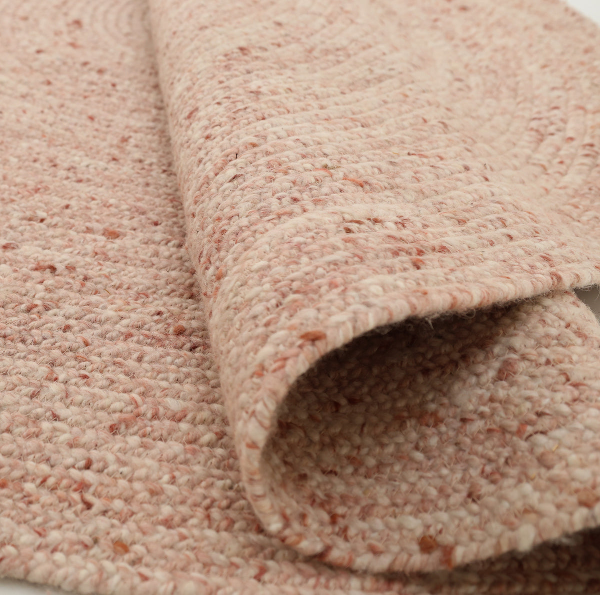 A close-up of the Soft Pink Braided Wool Area Rug Handmade Circular Rug- BR 099 by RAMSHA HOME reveals its textured, light pink surface, with part of the rug folded over to highlight its impressive thickness and intricate braided pattern.