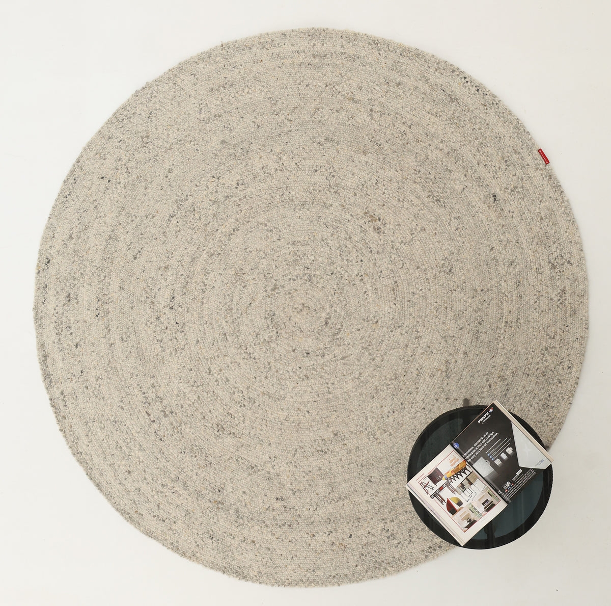 A round, light grey textured RAMSHA HOME Round Braided Wool Area Rug (Handmade Circular Rug- BR 100) sits on a white floor, complemented by a small black side table with an open magazine in the lower right corner. This handmade wool rug brings a soft, contemporary touch to any space.