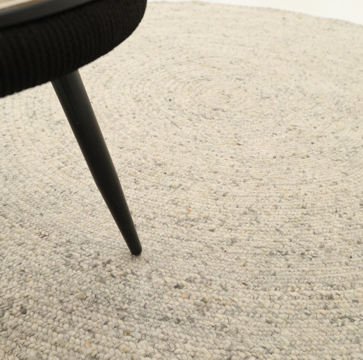 Close-up of a black chair leg resting on the textured, light gray surface of the RAMSHA HOME Round Braided Wool Area Rug Handmade Circular Rug- BR 100. The image highlights the rug’s intricate braided pattern and the modern, sleek design of the chair leg, beautifully showcasing the craftsmanship of this handmade round rug.