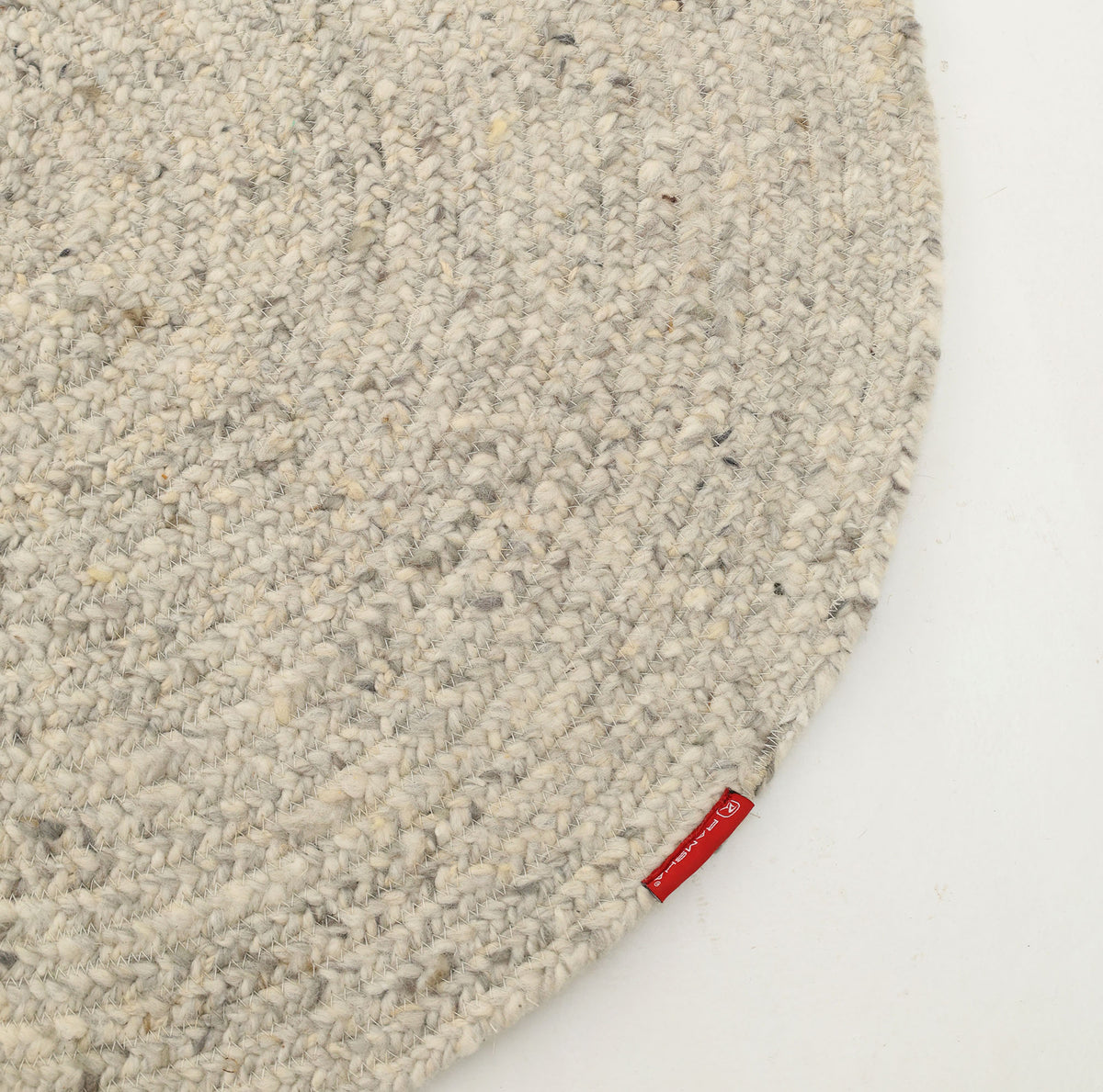 Presenting the RAMSHA HOME Round Braided Wool Area Rug Handmade Circular Rug- BR 100: a close-up of this handcrafted round rug showcases its light blush grey tone and intricate braided wool design, accented by a small red fabric tag on the edge, displayed on a white surface.