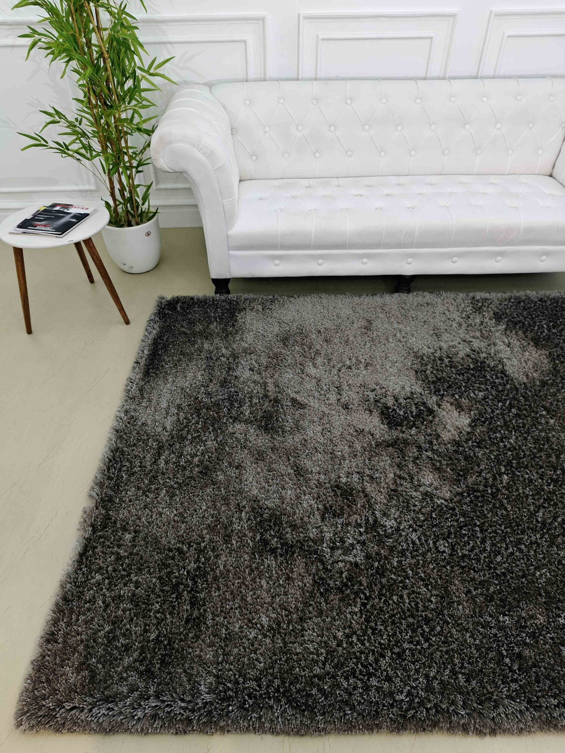 A modern living room features a white tufted sofa, a small round side table with magazines, a potted plant, and the RAMSHA HOME Grey Color Rectangle Shaped Shaggy Rug - SG 05 on a light floor for comfort and style.