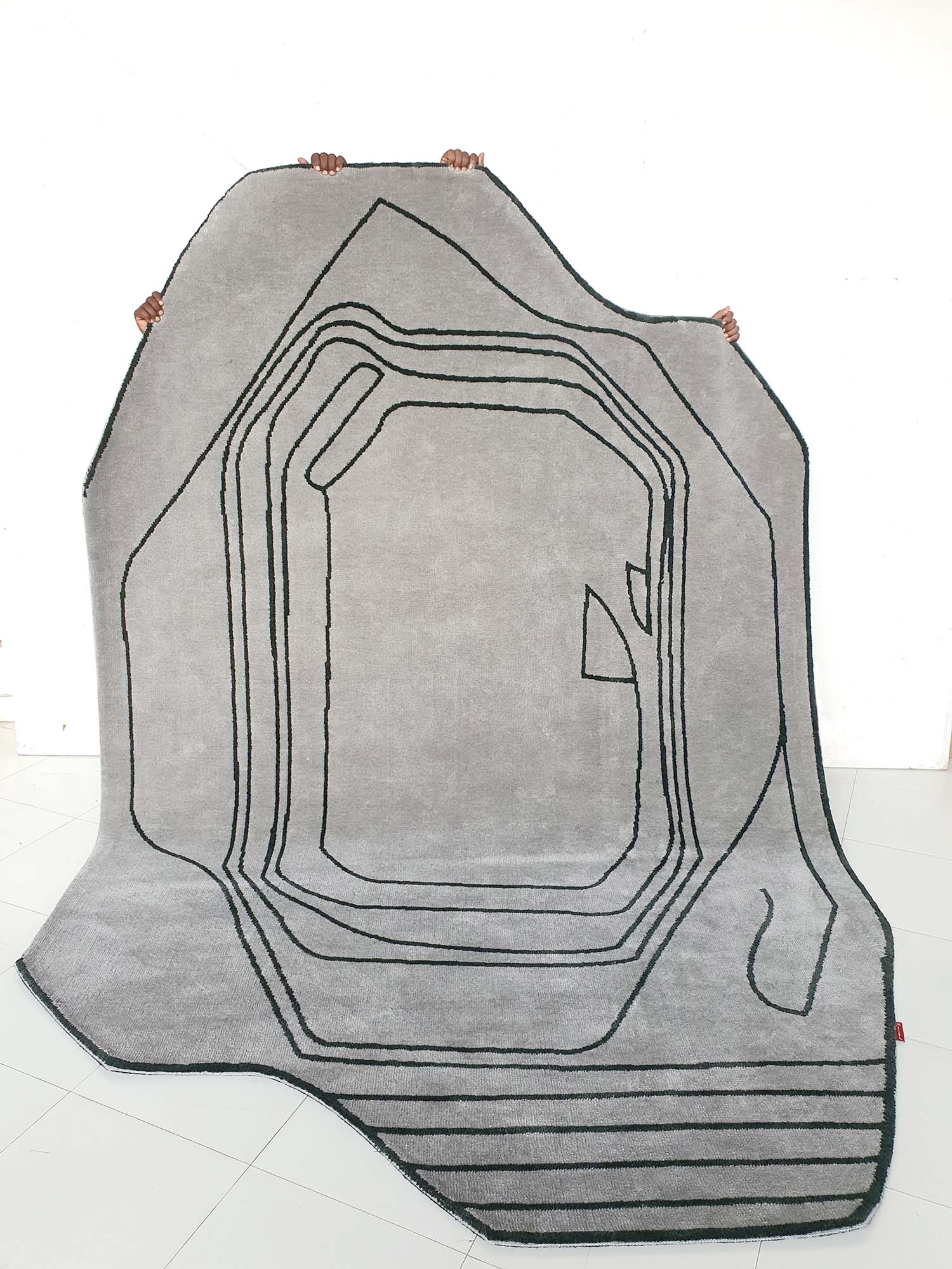 The RAMSHA HOME Shaped Rug Grey & Black Color Carpet (CMN-05), featuring a large irregular grey shape and black geometric lines, is held aloft by several hands with only the fingers visible against a plain white background.
