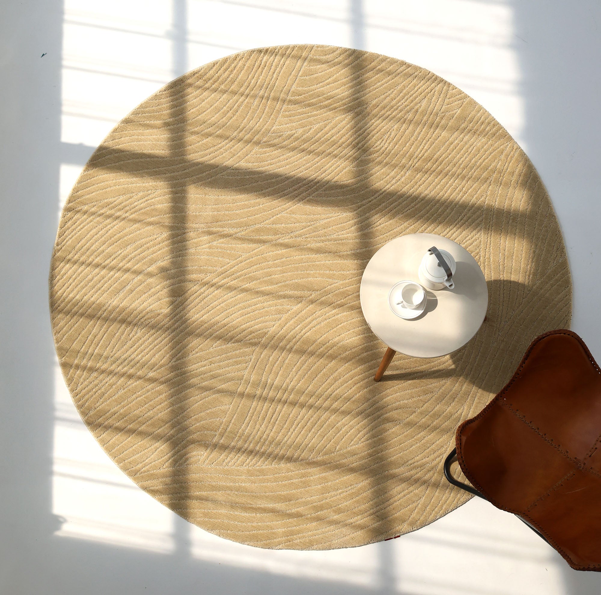 The RAMSHA HOME Modern Terrain Round Shaped Rug CMN 114 in beige is placed on a sunlit floor, alongside a small table with a teapot and cup. A brown leather chair peeks in, while shadows from the window enhance the tranquil setting.