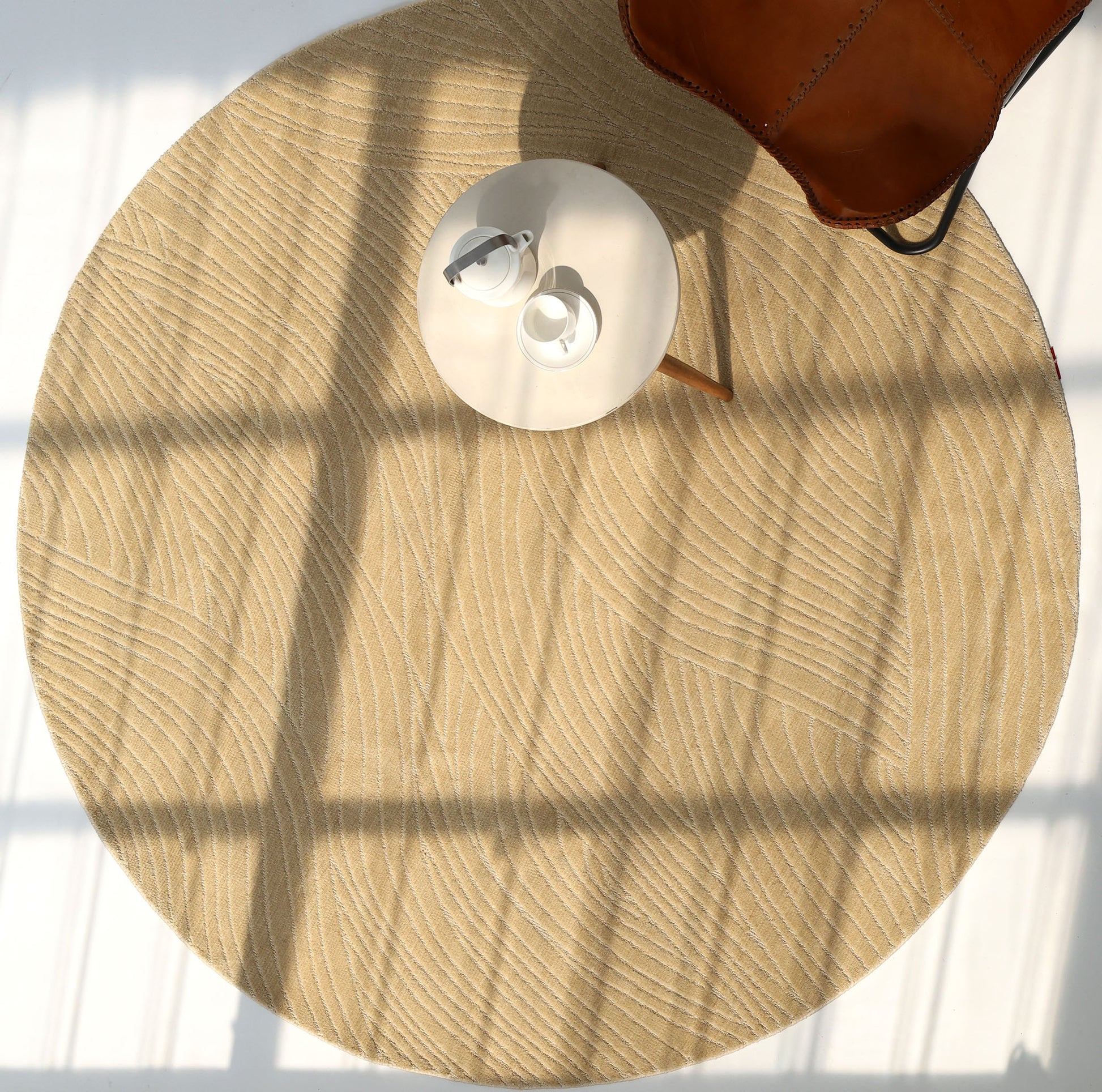 Sunlight streams through a window onto the RAMSHA HOME Modern Terrain Round Shaped Rug CMN 114, featuring beige wavy patterns. A small white table with a cup and saucer sits on the rug alongside a brown leather bag, all framed by window shadows.