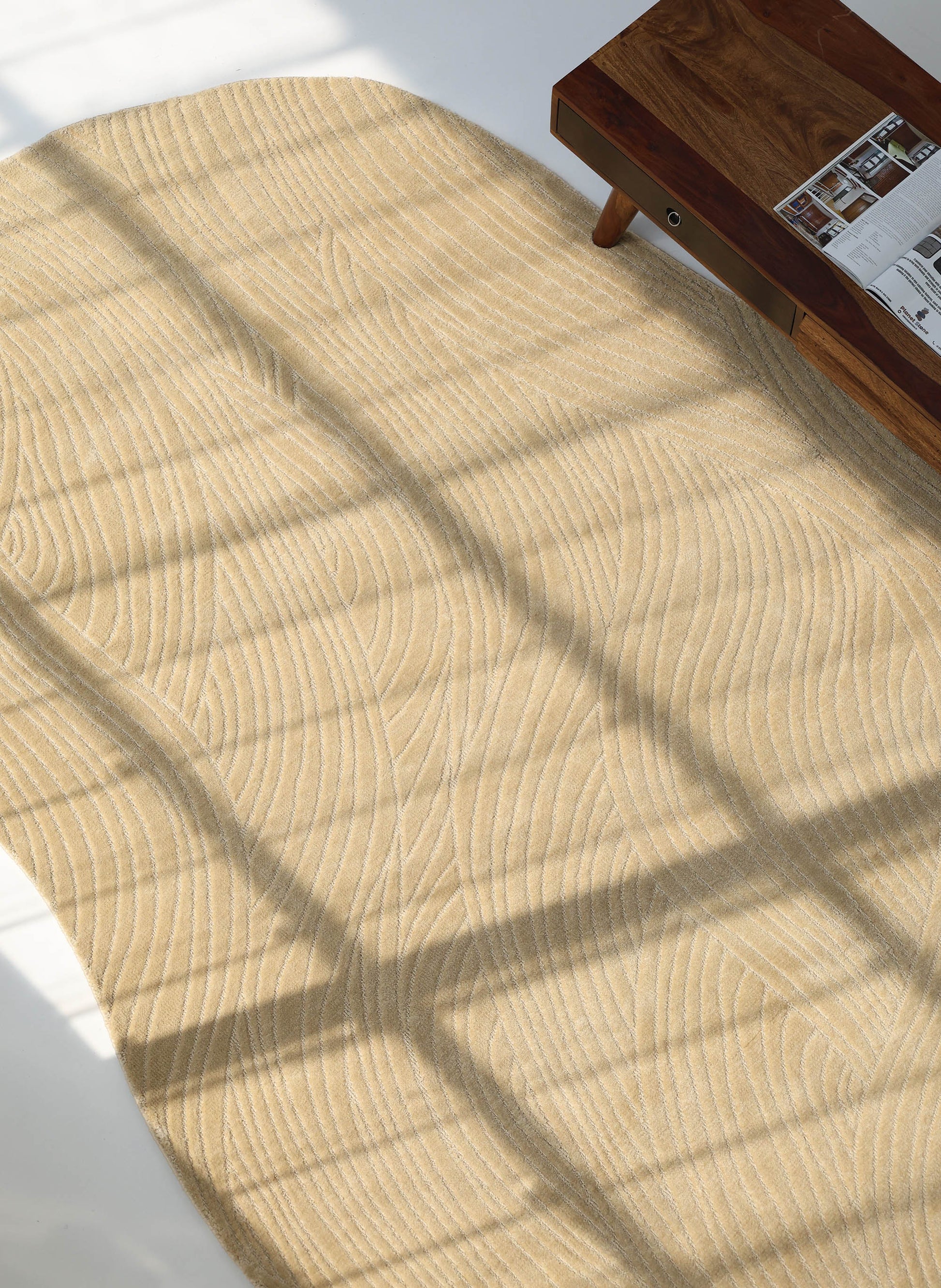 The RAMSHA HOME Modern Terrain Round Shaped Rug CMN 114, with wavy textured lines, sits in sunlight on a light floor as window shadows fall across it. A wooden table holding a magazine appears partially in the top right corner.