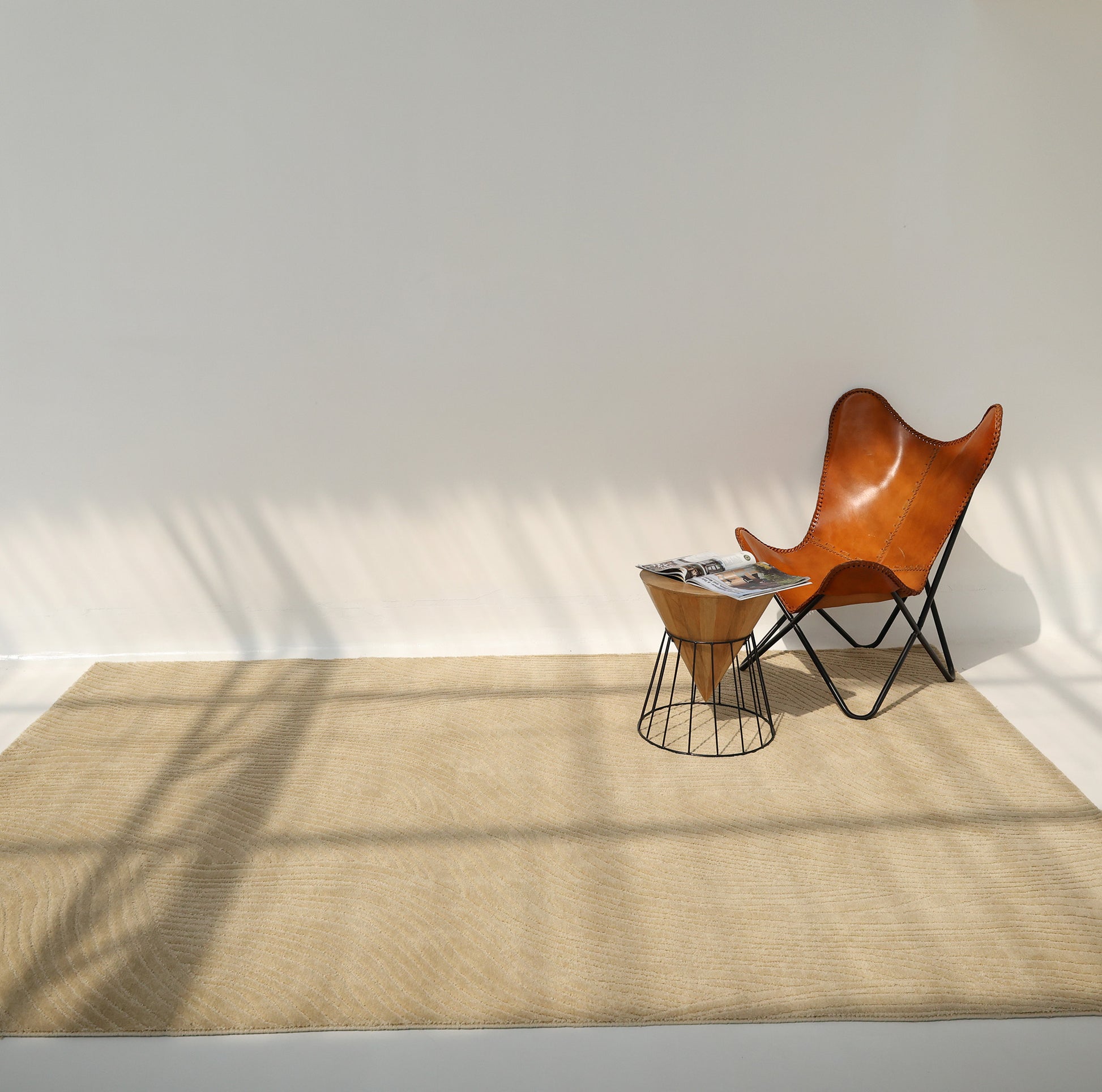 A minimalist room with a brown leather butterfly chair, a small wooden side table holding magazines, and the Modern Terrain Round Shaped Rug CMN 114 by RAMSHA HOME. Sunlight casts shadows across the light-colored wall and rug.