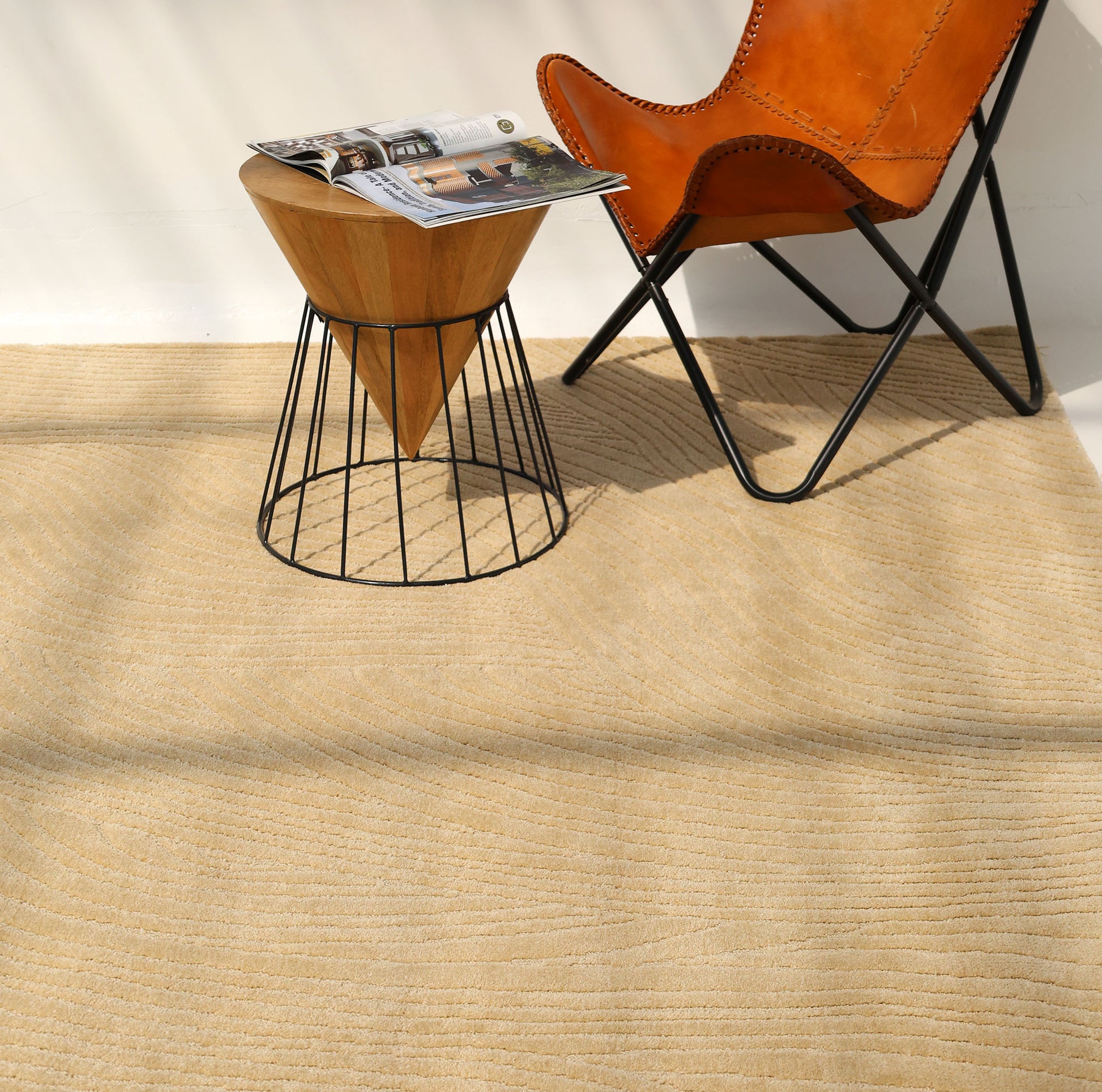 The RAMSHA HOME Modern Terrain Round Shaped Rug CMN 114 in tan with subtle wave patterns sits under a brown leather chair and a wood-metal side table with an open magazine, all illuminated by natural sunlight.
