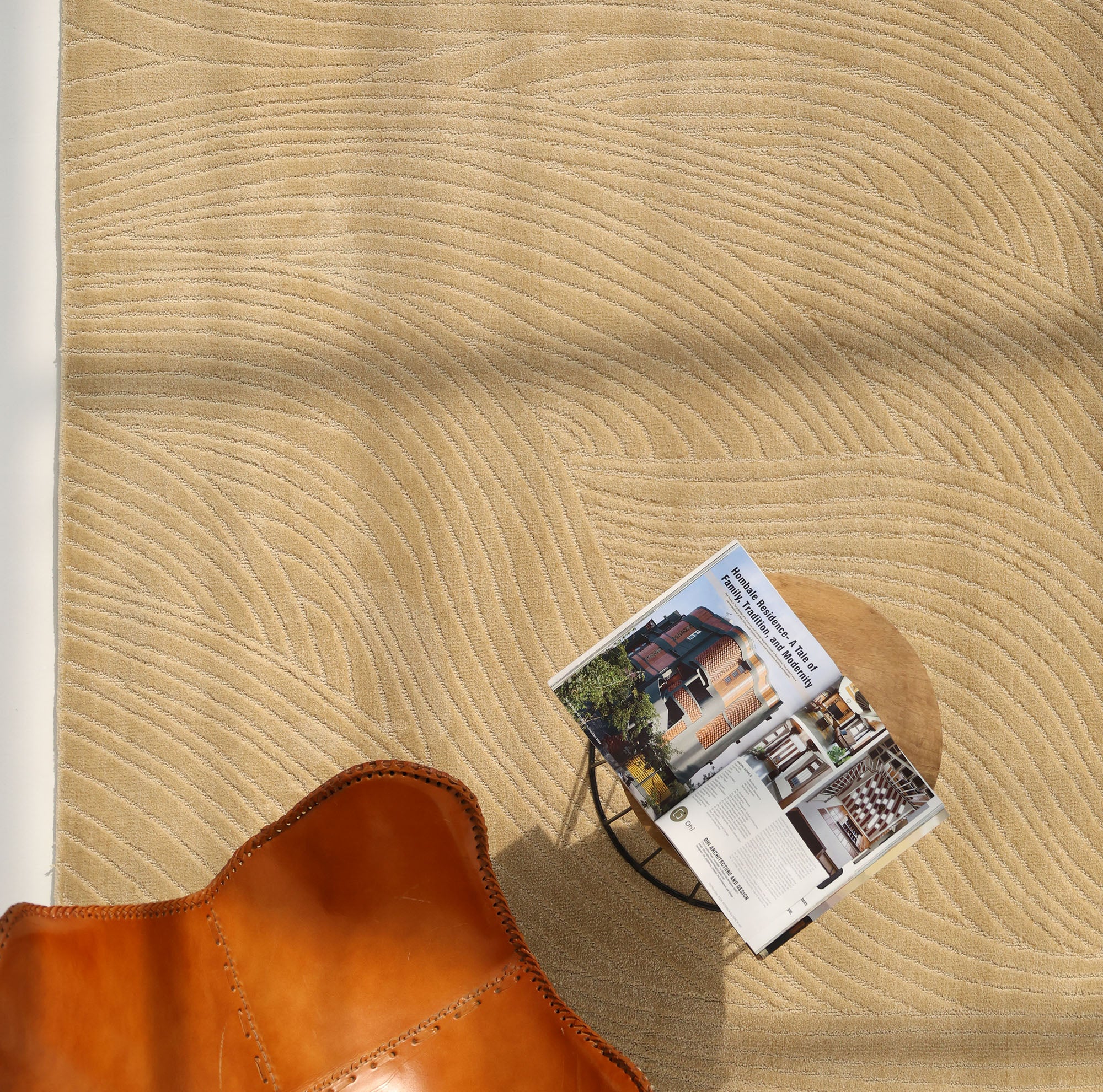The RAMSHA HOME Modern Terrain Round Shaped Rug CMN 114, featuring tan wavy textures, is paired with a brown leather chair and a small table holding an open magazine. Sunlight streams in, casting soft shadows across the space.