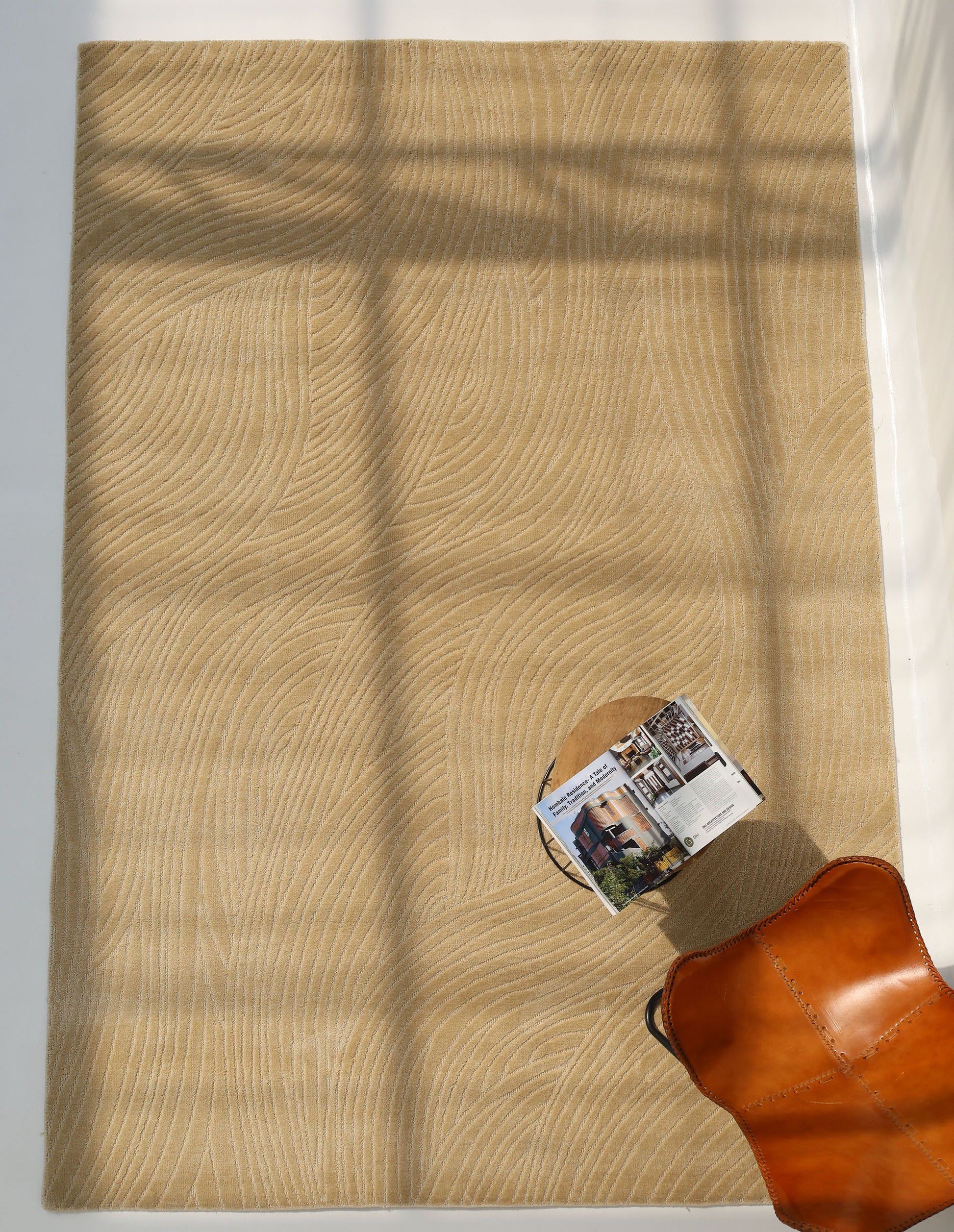 The RAMSHA HOME Modern Terrain Round Shaped Rug CMN 114, featuring a tan wavy textured design, sits on a white floor with sunlight casting window grid shadows. A brown leather chair and a small round table with an open magazine are nearby.