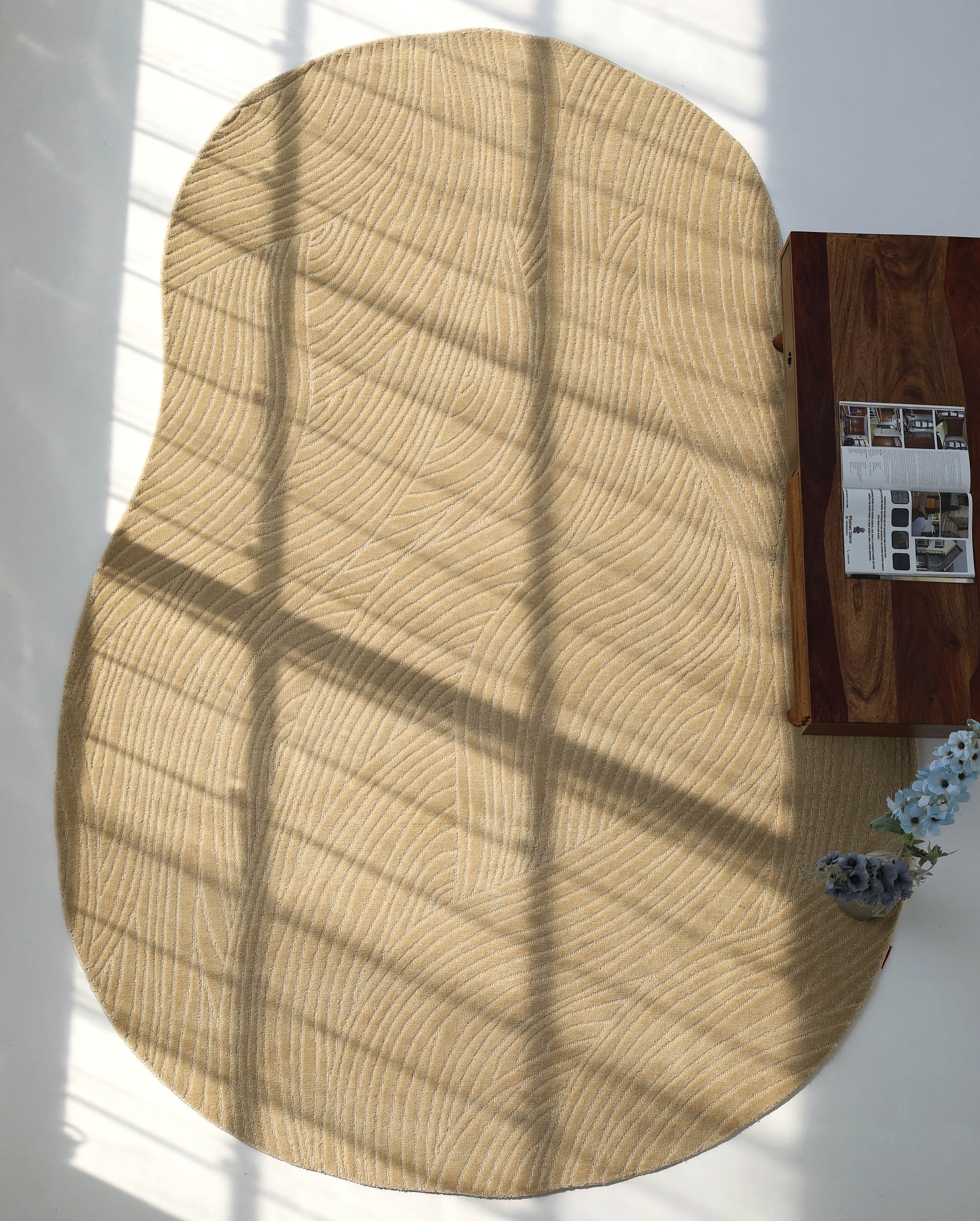 The Modern Terrain Round Shaped Rug CMN 114 by RAMSHA HOME, featuring wavy abstract patterns in beige, is placed by a wooden table with a magazine and blue flowers. Window pane shadows gently fall across the rug.