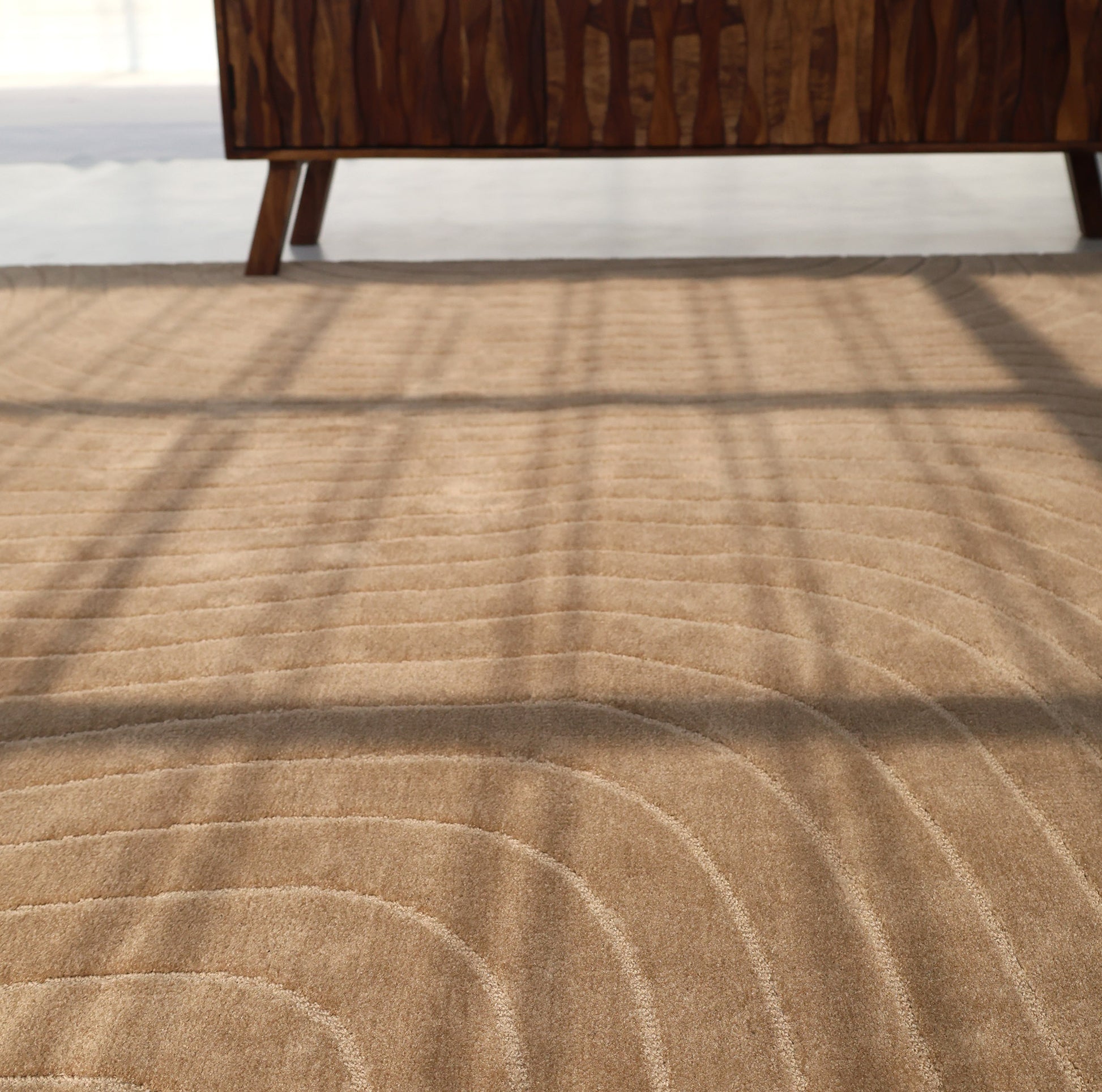 Close-up of RAMSHA HOME’s Earth Lines Contemporary Area Shaped Rug CMN 115, featuring light brown tones and subtle curved lines, with sunlight creating grid-like shadows and a wooden cabinet partially visible in the background.