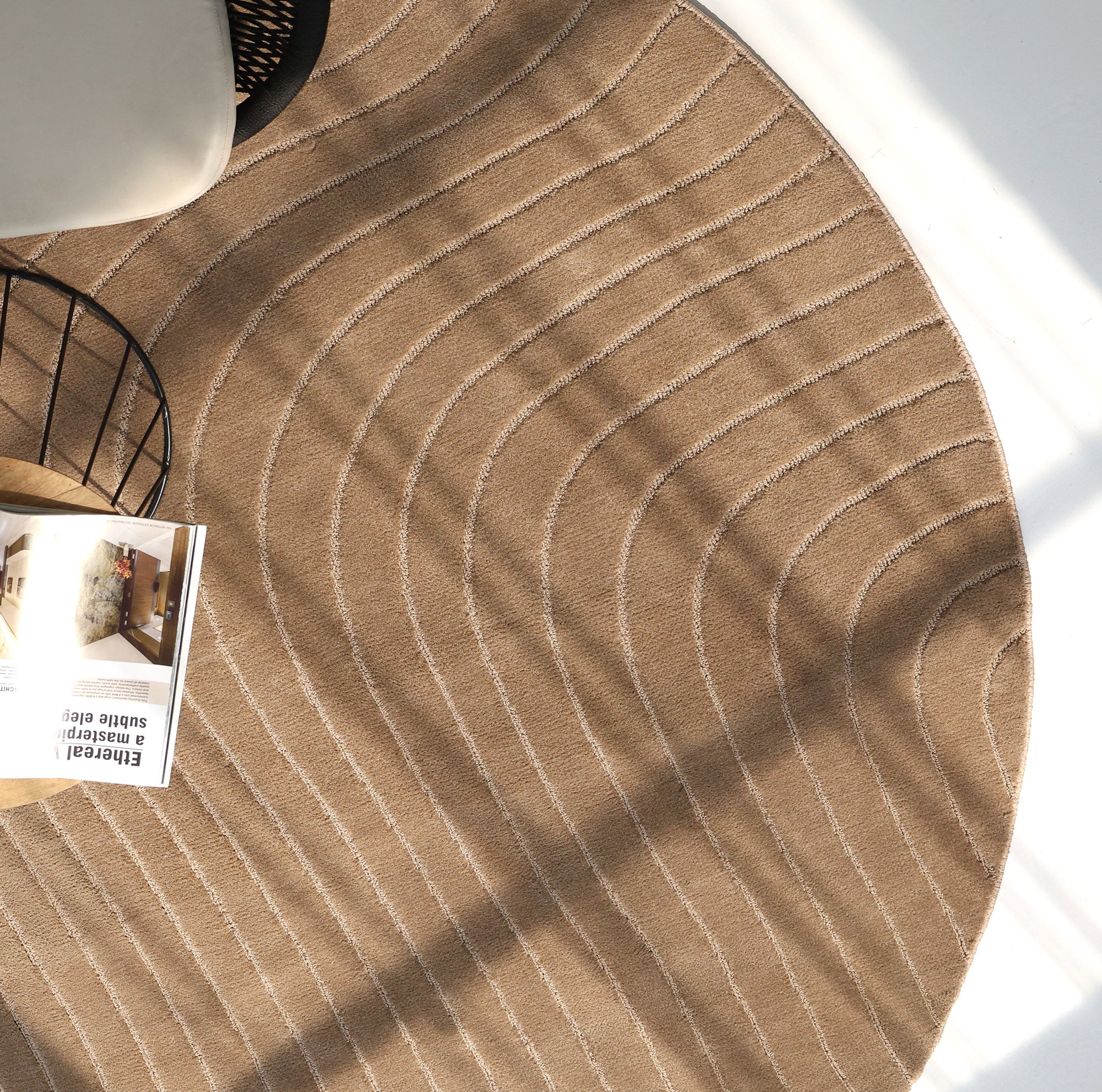 The Earth Lines Contemporary Area Shaped Rug CMN 115 by RAMSHA HOME, featuring a round beige design with curving white lines, is partly visible on a sunlit floor near a wire-frame side table and a cream-colored chair.