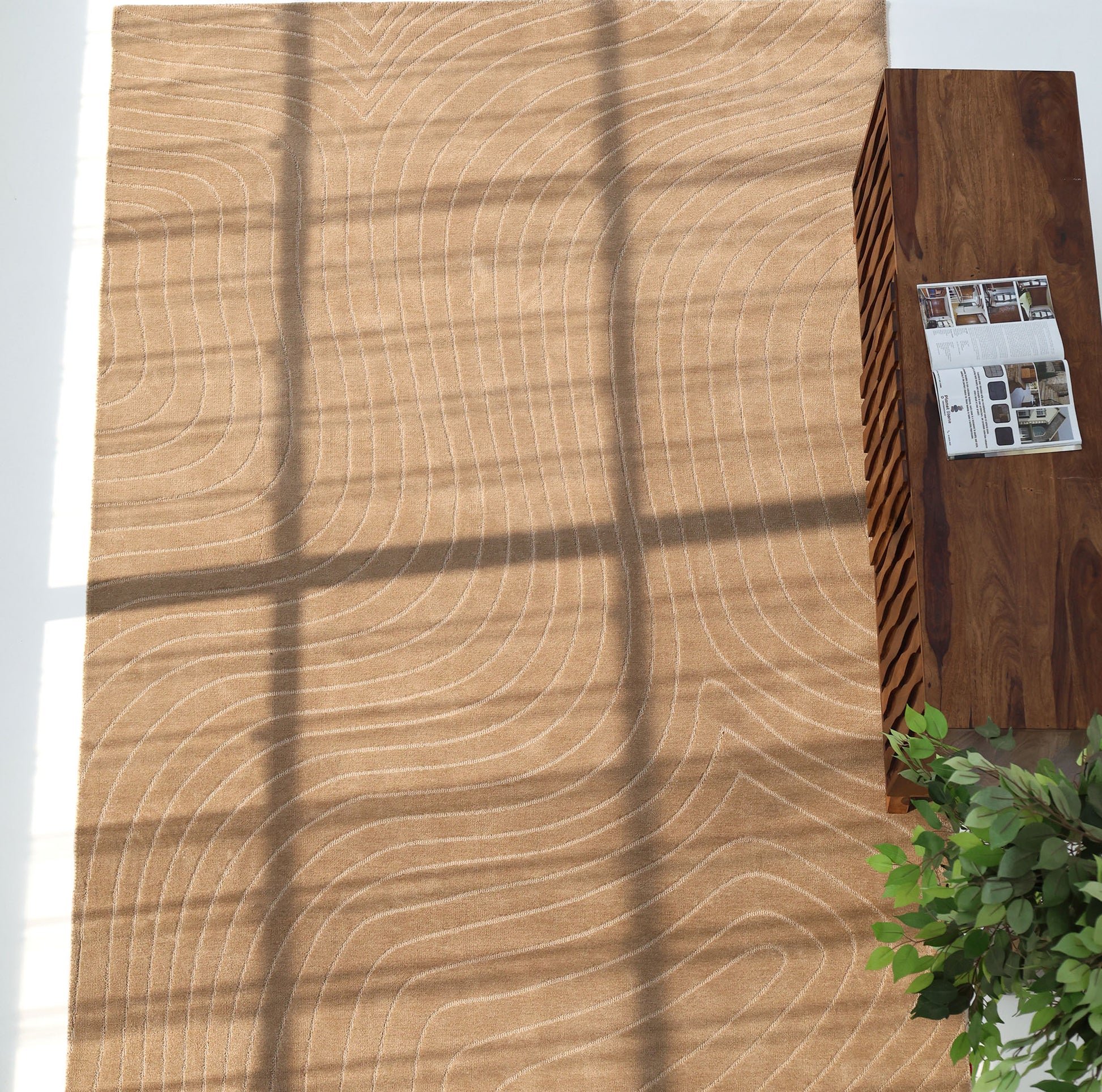 The RAMSHA HOME Earth Lines Contemporary Area Shaped Rug CMN 115 features a light brown modern pattern with curved white lines, sunlight casting windowpane shadows, and is paired with a wooden table, magazines, and a green plant in the corner.