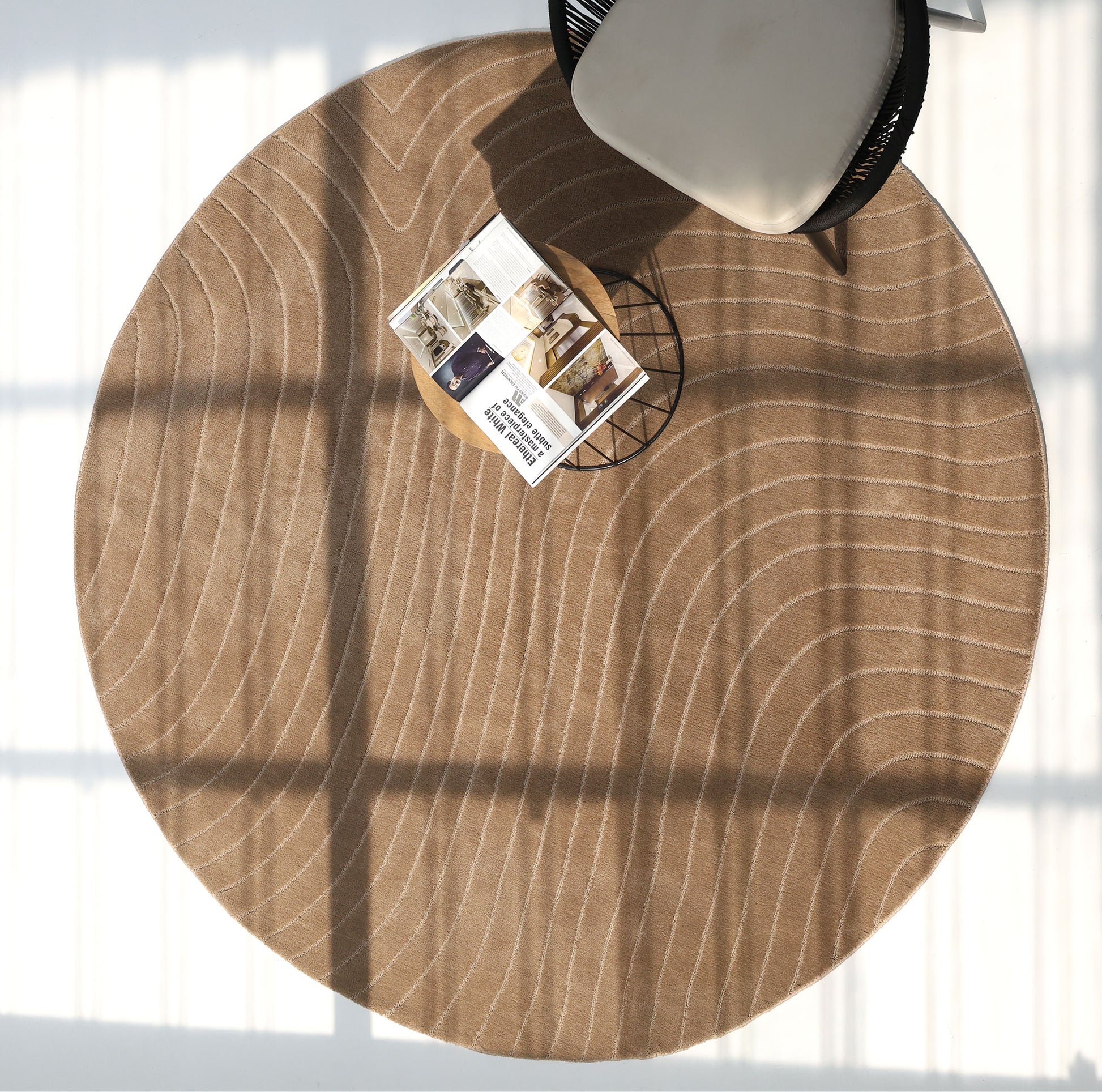 The RAMSHA HOME Earth Lines Contemporary Area Shaped Rug CMN 115, featuring a modern beige design with curved white lines, lies on the floor. A wire-frame table with an open magazine is nearby, illuminated by window-cast sunlight.