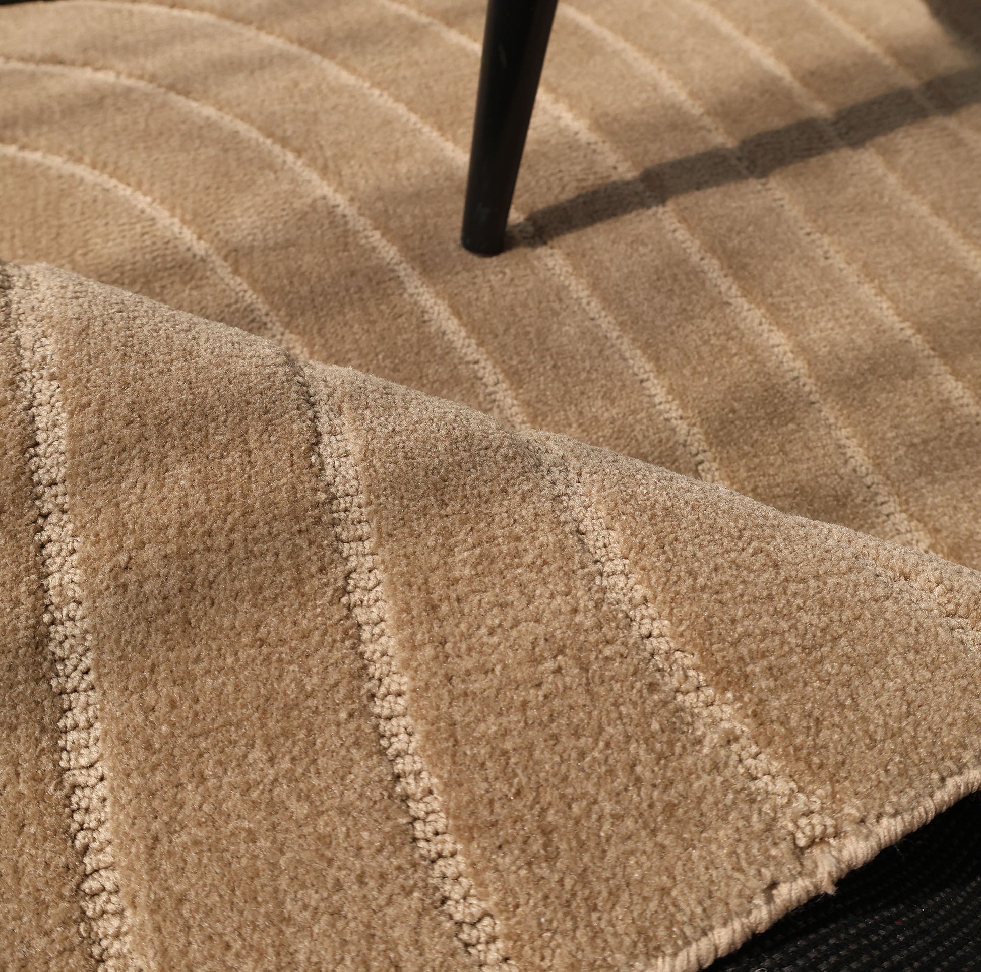 A close-up of the RAMSHA HOME Earth Lines Contemporary Area Shaped Rug CMN 115 in beige, with curved, raised lines; part of the rug is lifted to show a black surface beneath, and a thin black table or chair leg rests on its modern design.