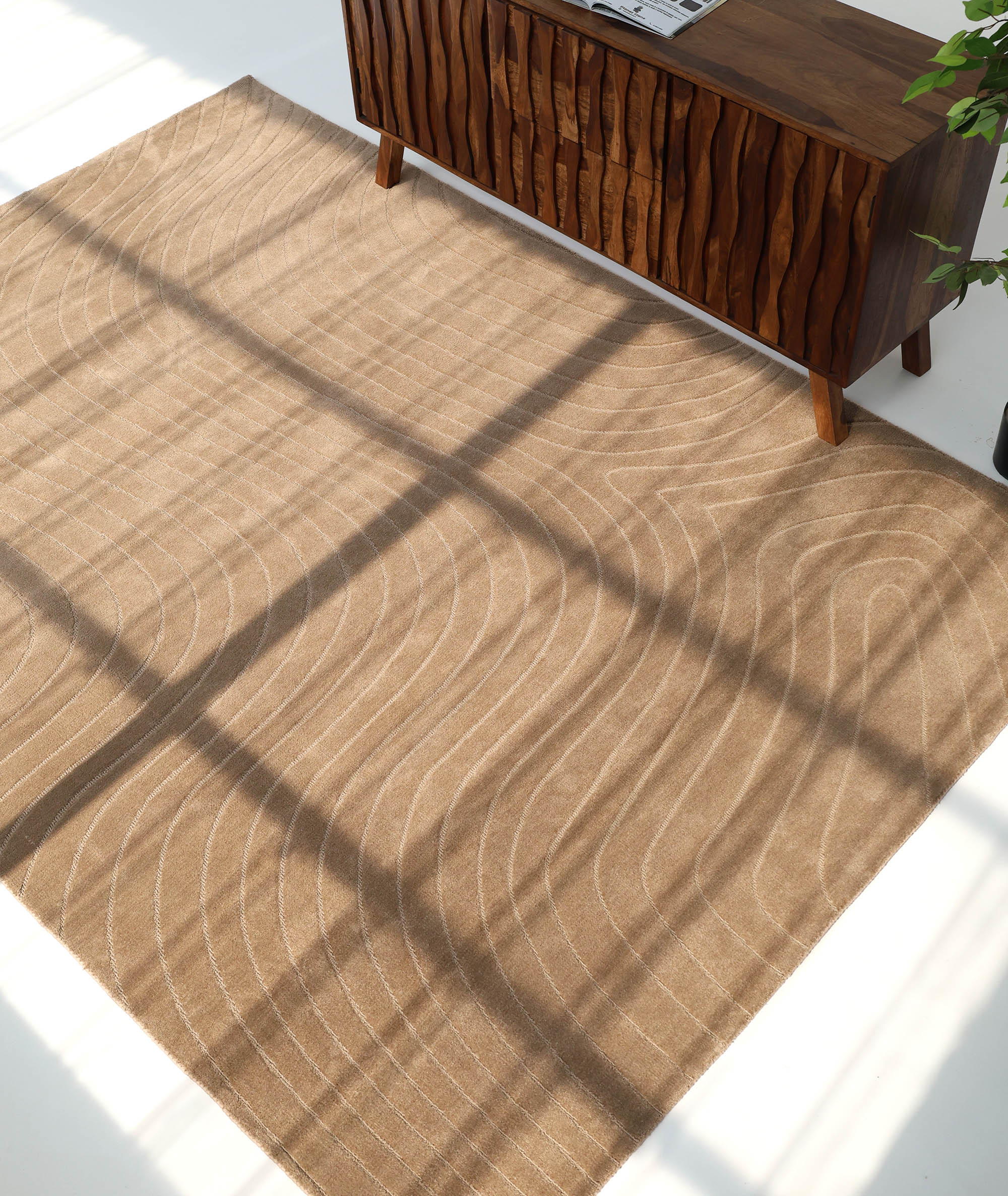 The RAMSHA HOME Earth Lines Contemporary Area Shaped Rug CMN 115, in light brown with modern wavy patterns, is placed before a wooden cabinet as sunlight through a window casts a grid-like shadow across the rug and floor.