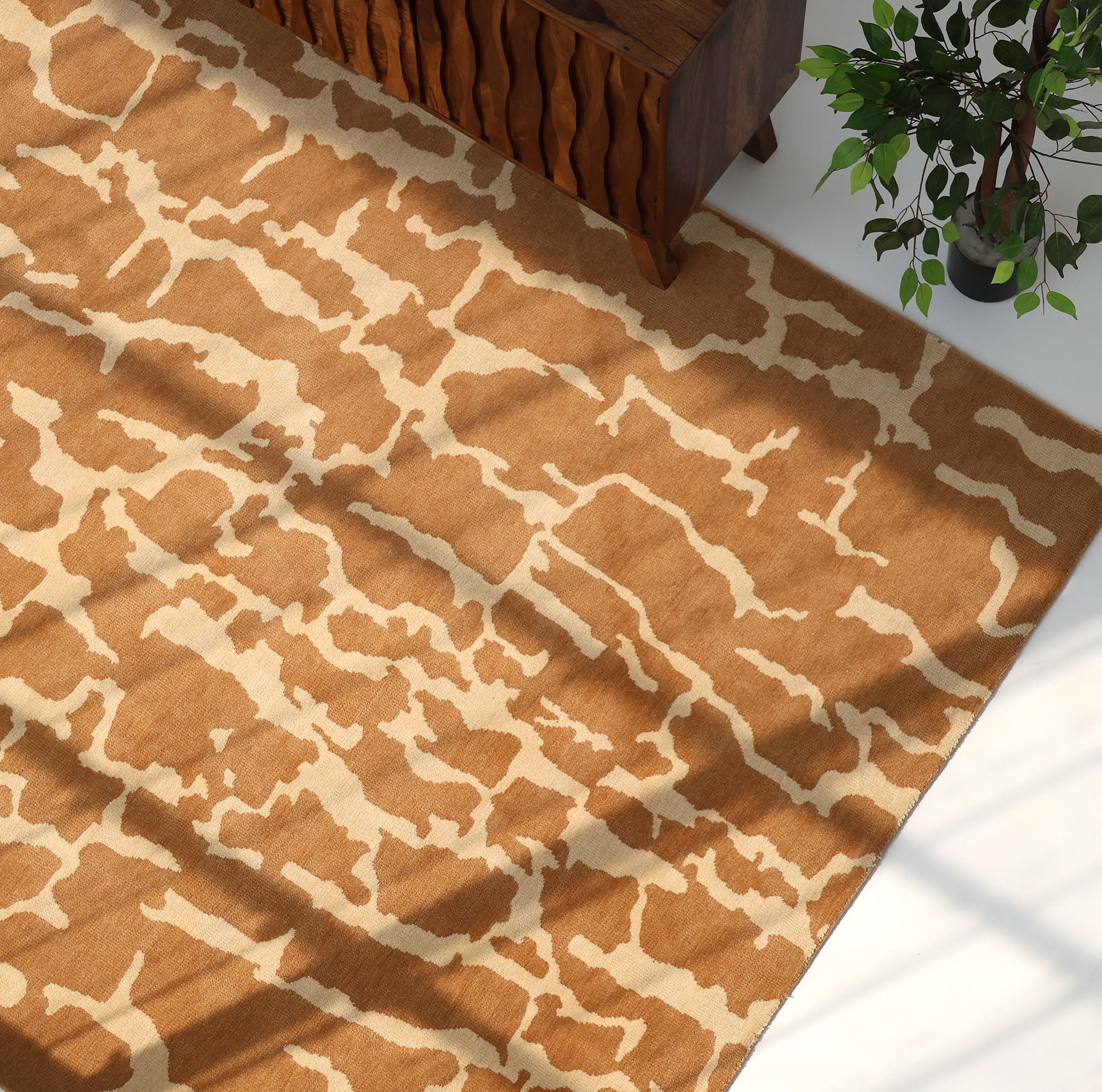 The RAMSHA HOME Natural Drift Round Area Rug CMN 116, featuring a modern brown and beige abstract pattern, sits partly beneath a wooden cabinet beside a leafy plant as sunlight gently highlights its handcrafted design.