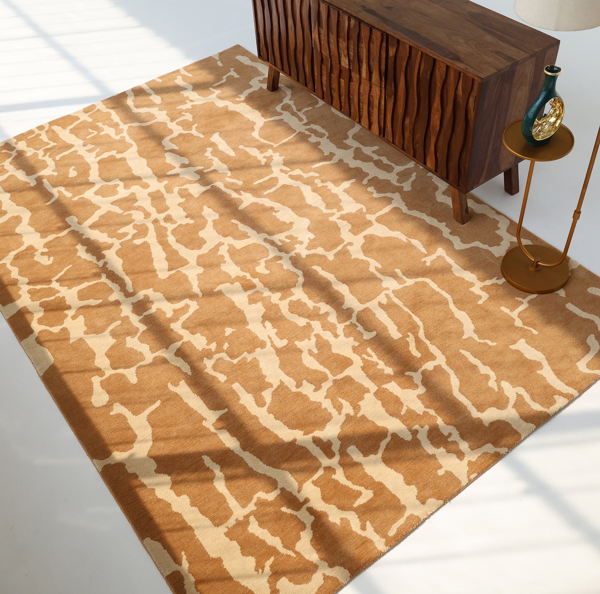 The RAMSHA HOME Natural Drift Round Area Rug CMN 116, featuring a tan and cream abstract design, is placed beside a wavy wooden credenza, a round side table with a gold lamp, and a vase as sunlight casts shadows across the room.