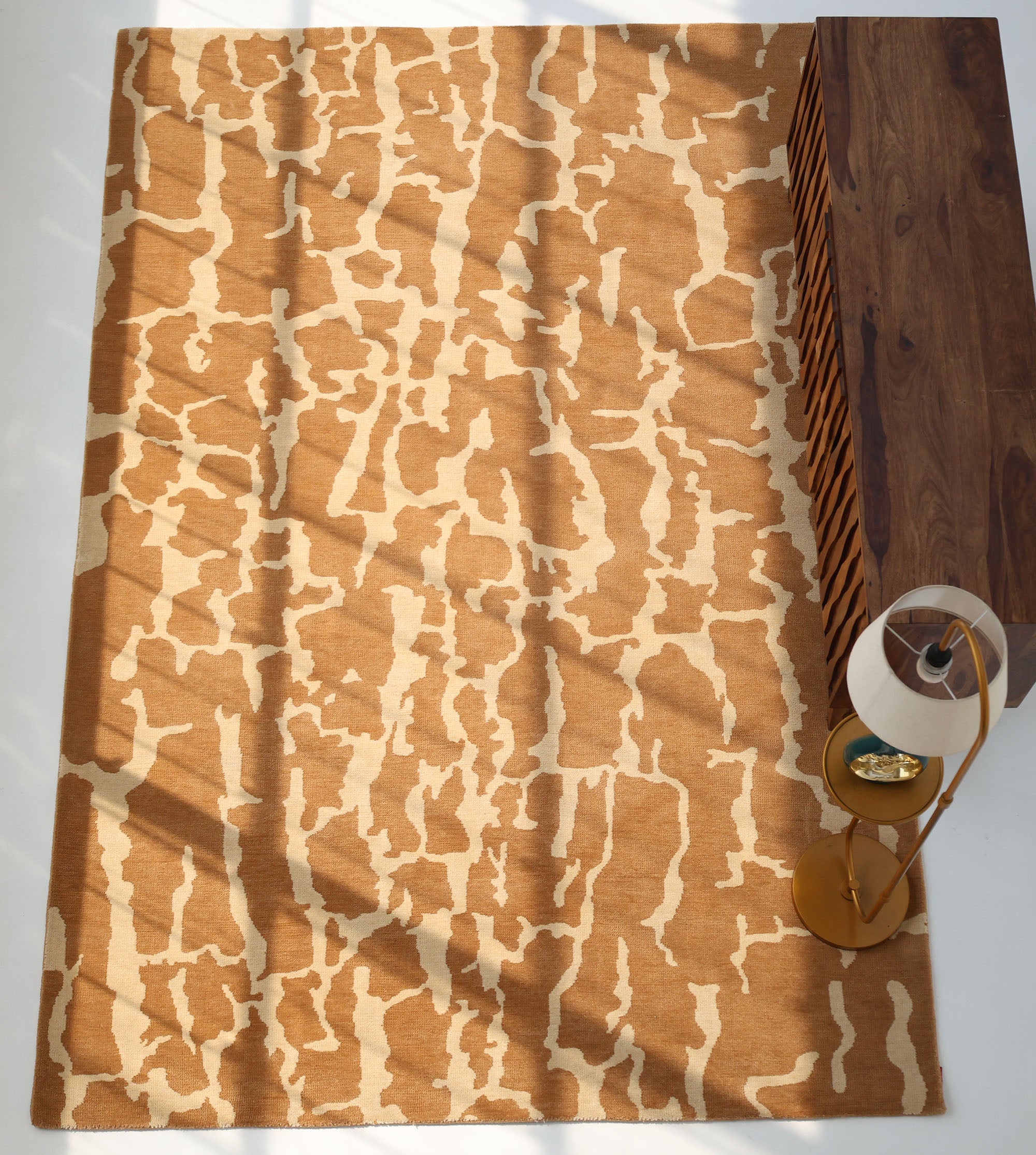 The RAMSHA HOME Natural Drift Round Area Rug CMN 116 features an abstract tan and beige design. Positioned near a wooden cabinet and a gold-based floor lamp, sunlight casts shadows across the stylish round rug.